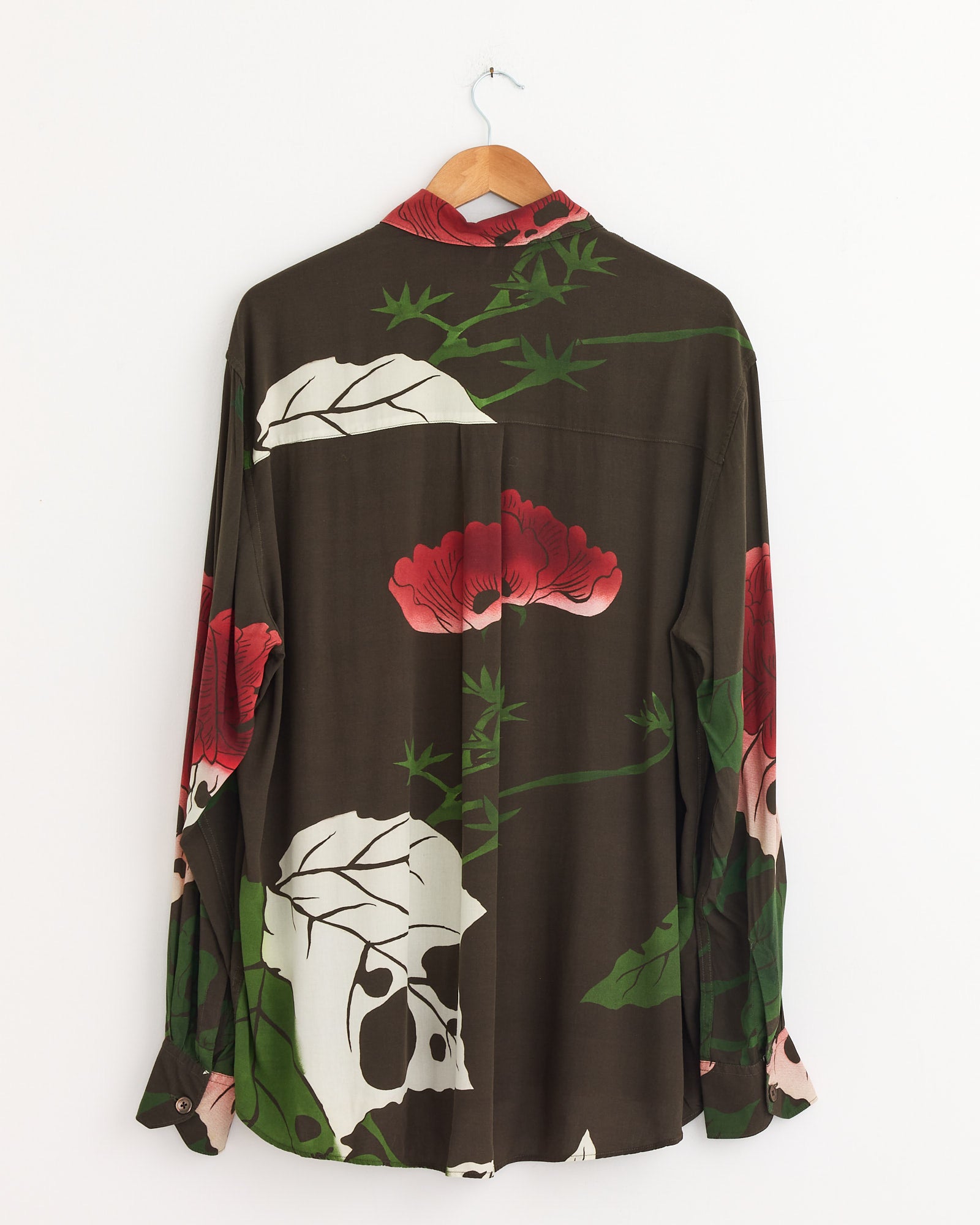 The Above Shirt in Hell Of Ween Print by Our Legacy, a dark long-sleeve Italian rayon button-up with a red, white, and green floral pattern, hangs from a wooden hanger against a white wall, shown from the back.