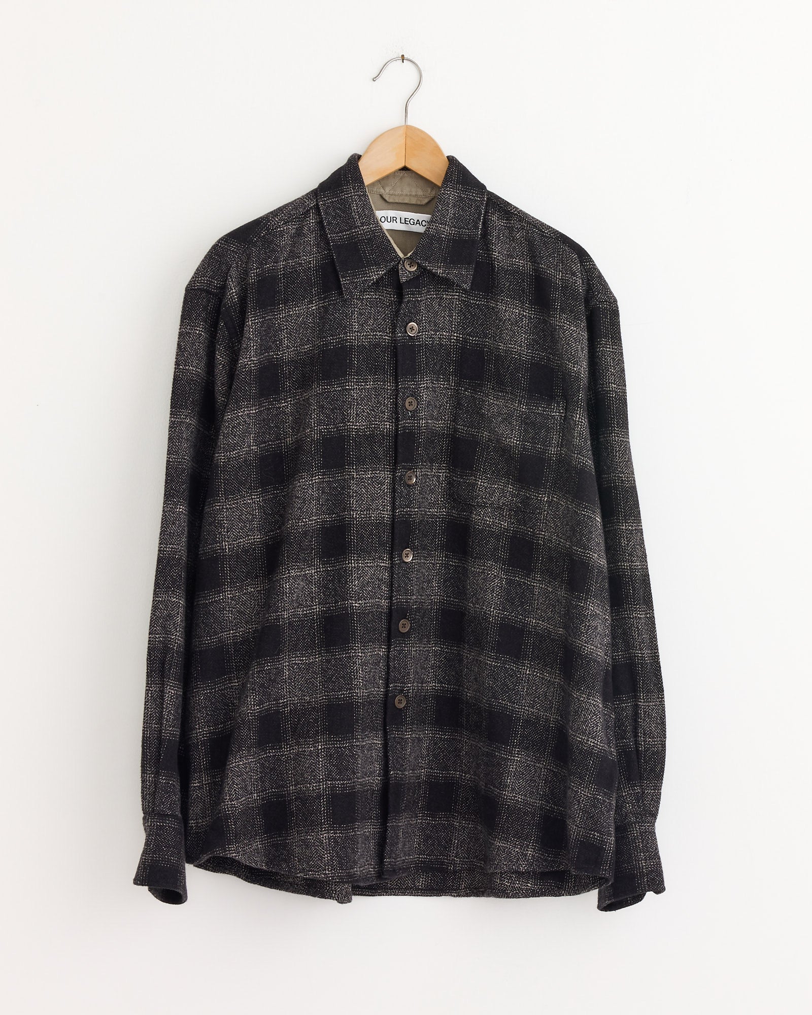 The Our Legacy Above Shirt in Oakland Low Twist Check, a black and gray plaid button-up with long sleeves made from Japanese cotton, hangs on a wooden hanger against a plain white wall.