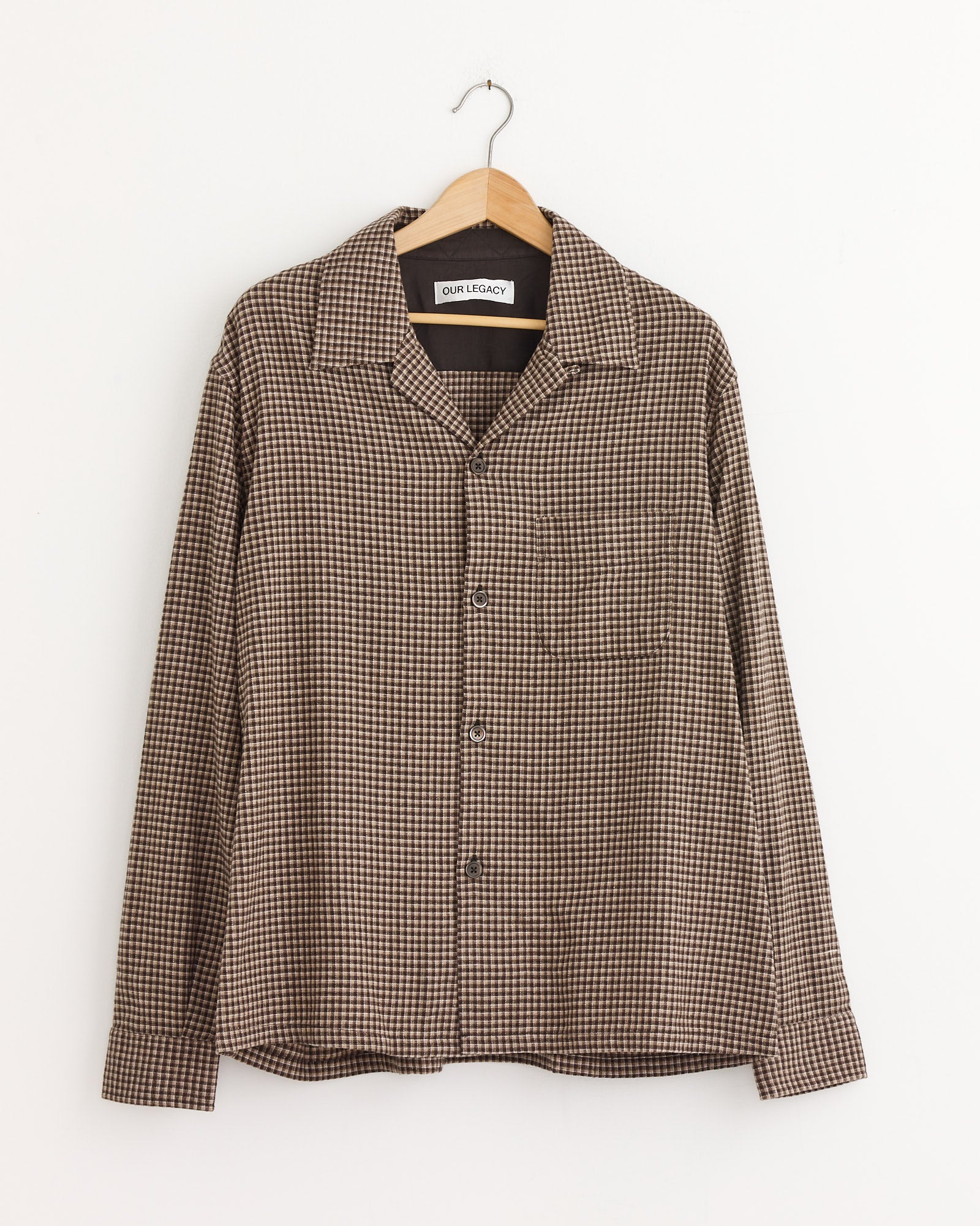 The Our Legacy Heusen Shirt in Endino Flannel Check, featuring a collar, chest pocket, and boxy fit, hangs on a wooden hanger against a white wall.