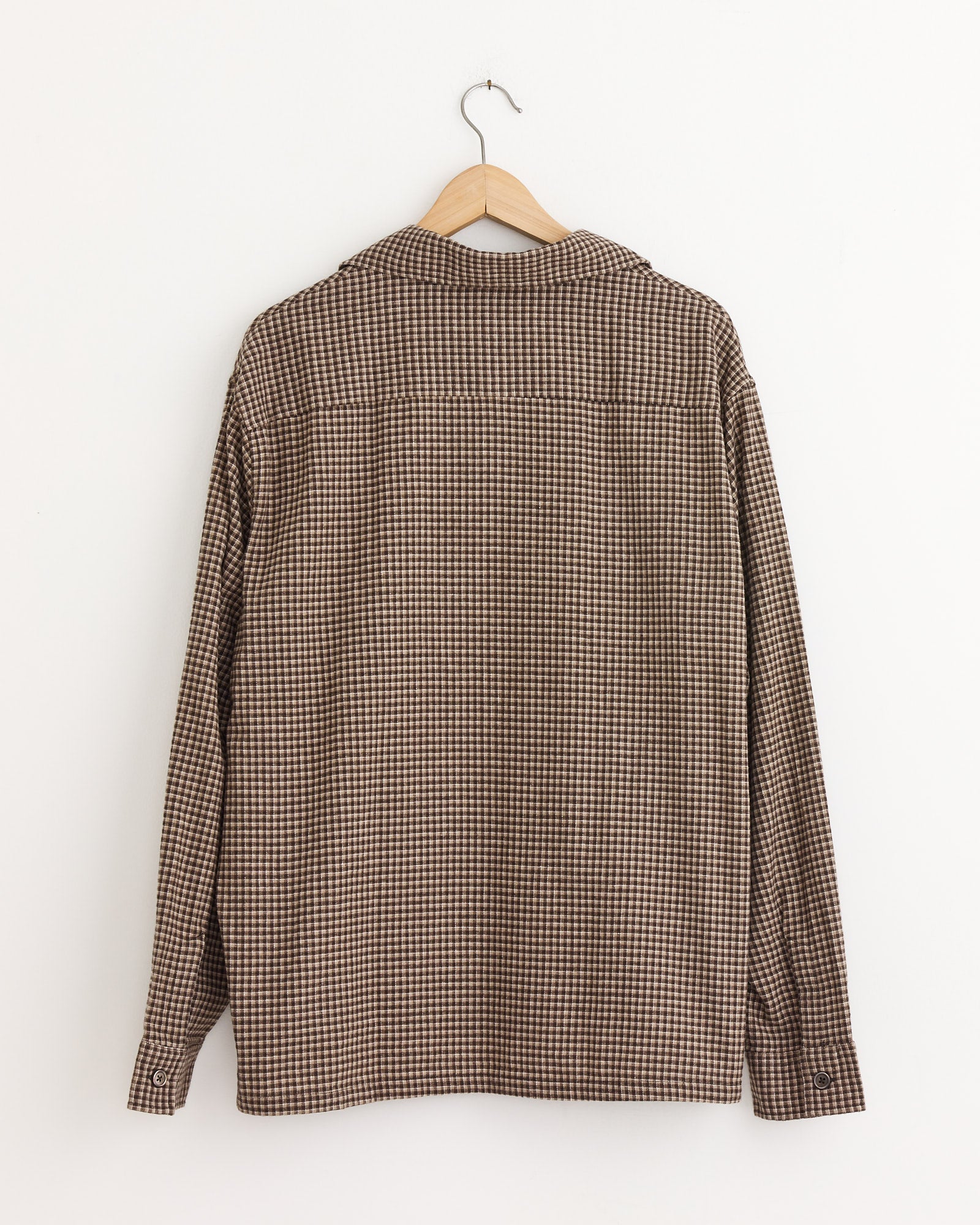 The Our Legacy Heusen Shirt in Endino Flannel Check, featuring a boxy fit in brown and beige checks, is displayed from the back on a wooden hanger against a plain white wall.