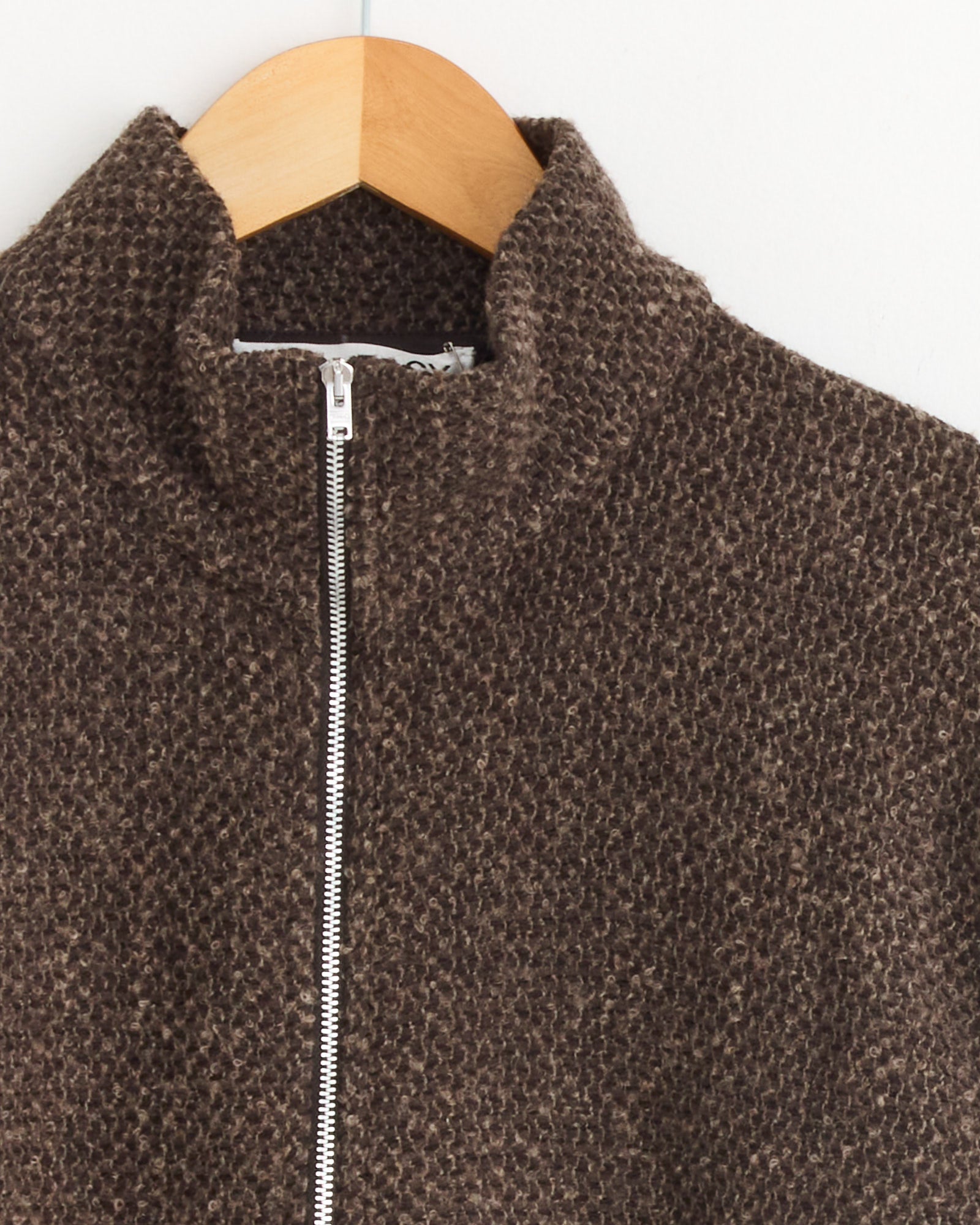 A close-up of the Our Legacy Shrunken Full Zip Polo in Brown Sparse, featuring textured Italian fabric, a high collar, and silver zipper, displayed on a wooden hanger against a white wall.