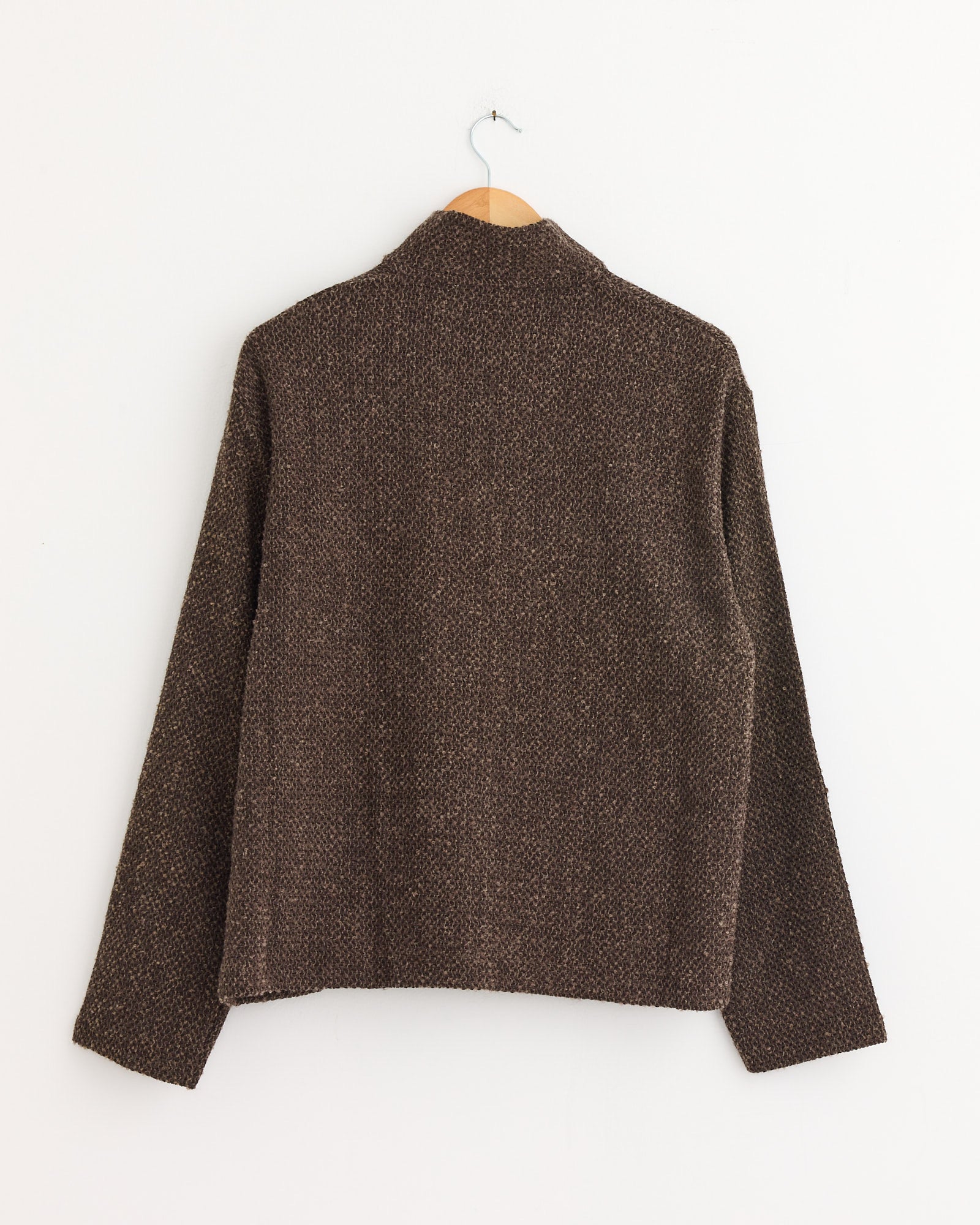 The Shrunken Full Zip Polo in Brown Sparse by Our Legacy, featuring textured Italian fabric and long sleeves, hangs on a wooden hanger against a white wall, displayed from the back.