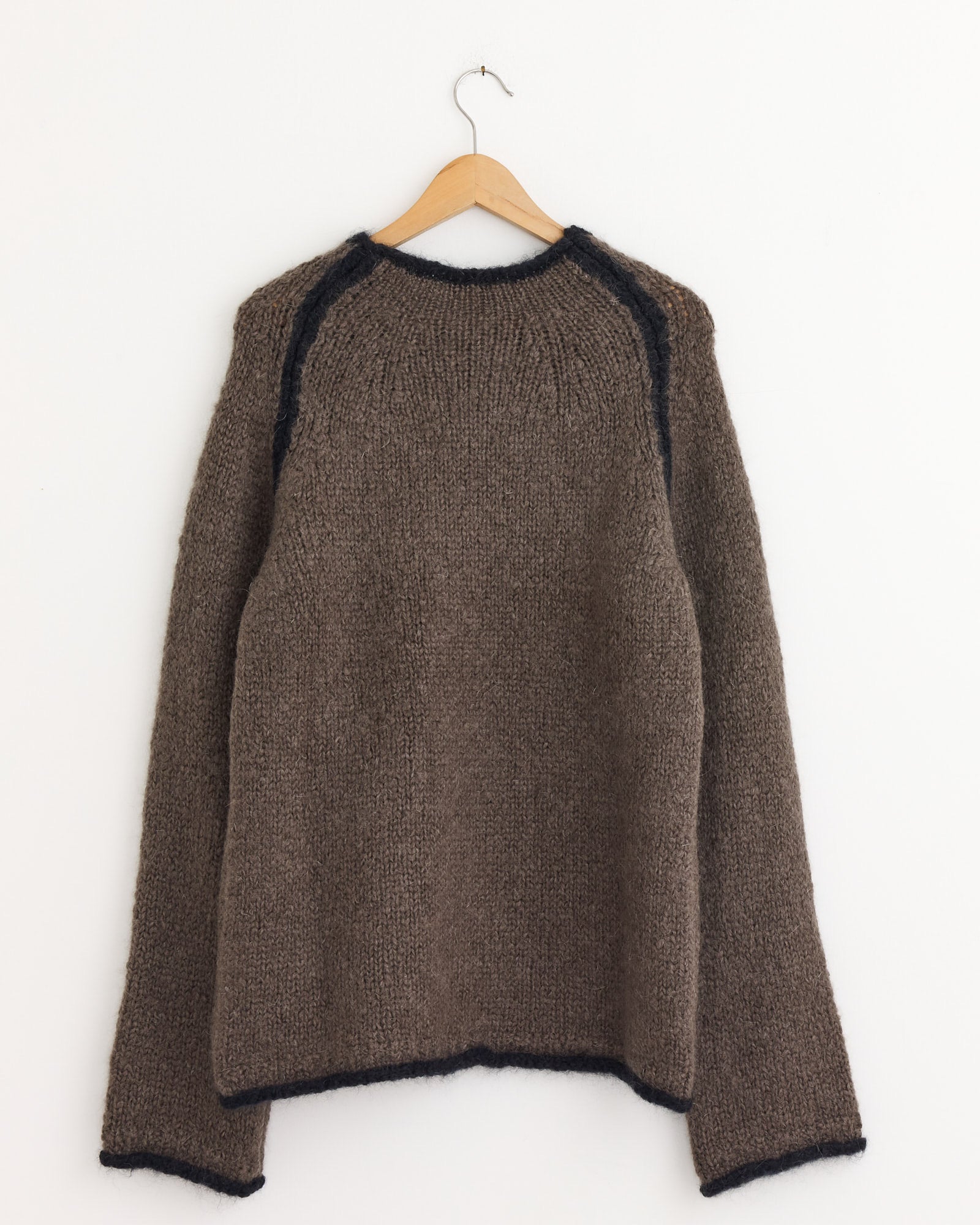 The Our Legacy Rider Roundneck Sweater in Steel—a men's relaxed fit, long-sleeved knitted sweater with dark trim on the neck, shoulder seams, cuffs, and hem—hangs on a wooden hanger against a plain white wall.