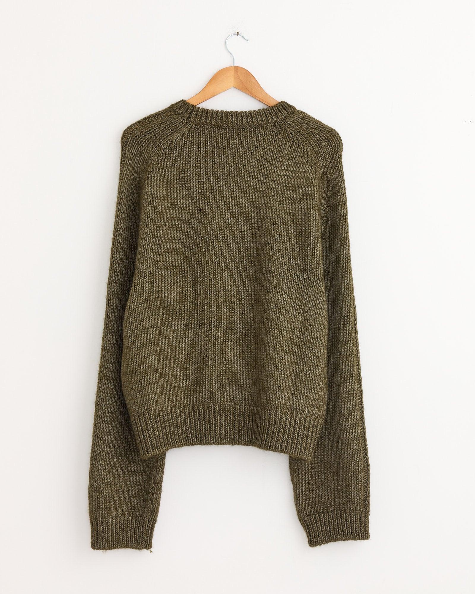 A True Roundneck Sweater in Flush Green by Our Legacy, crafted from Italian wool silk with long sleeves, hangs on a wooden hanger against a plain white wall.