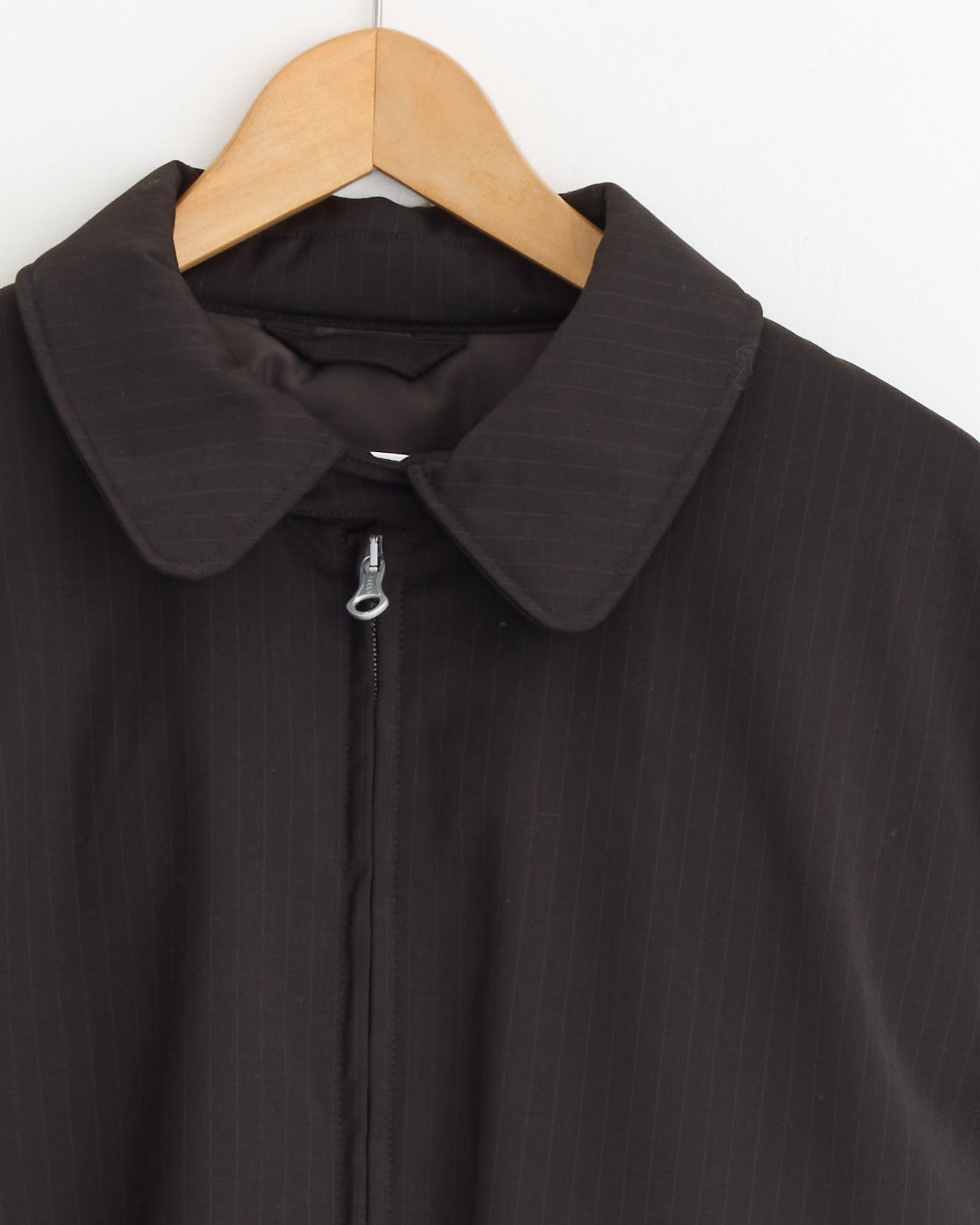 The Avenue Puffa Jacket in Churchill Stripe by Our Legacy is a dark brown pinstripe men's boxy fit outerwear with a wide collar and zipper, displayed on a wooden hanger against a white background.