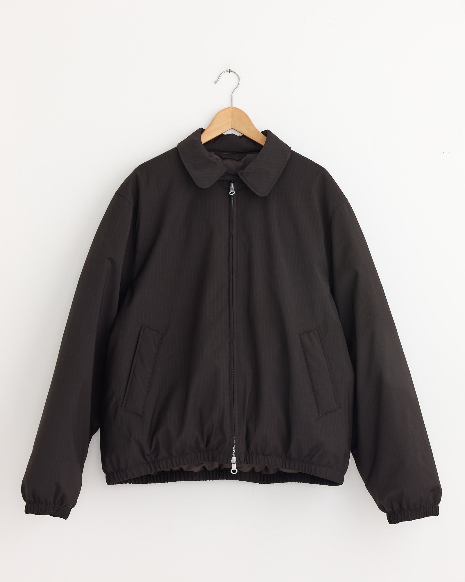 The Avenue Puffa Jacket in Churchill Stripe by Our Legacy features a dark brown zip-up design with a collar, two slanted front pockets, elastic cuffs and hem, displayed on a wooden hanger against a plain white wall.
