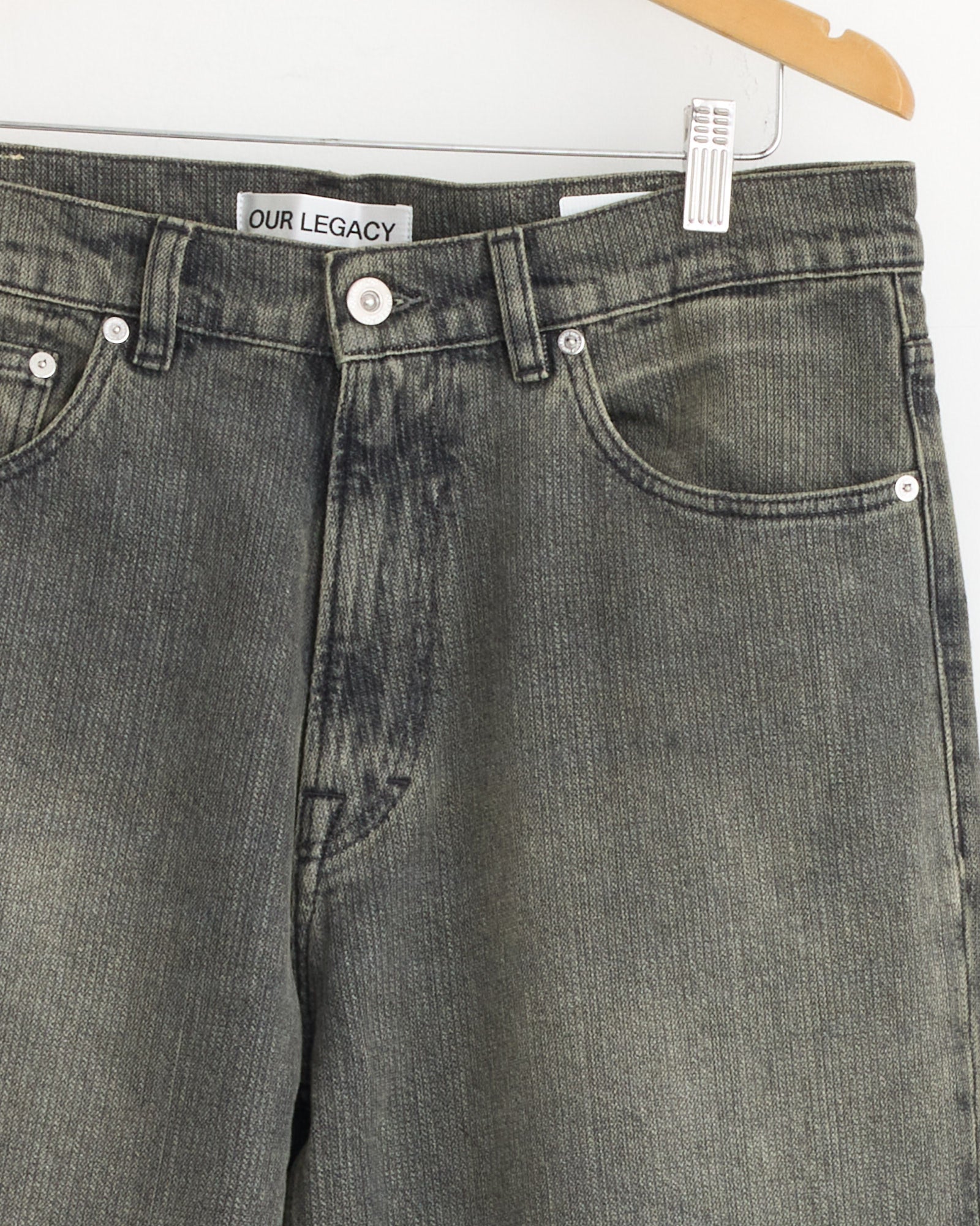 Close-up of Our Legacy Third Cut Jeans in Ash Dust, displayed on a wooden hanger. Features include visible pockets, belt loops, white buttons, and an OUR LEGACY label inside the waistband.
