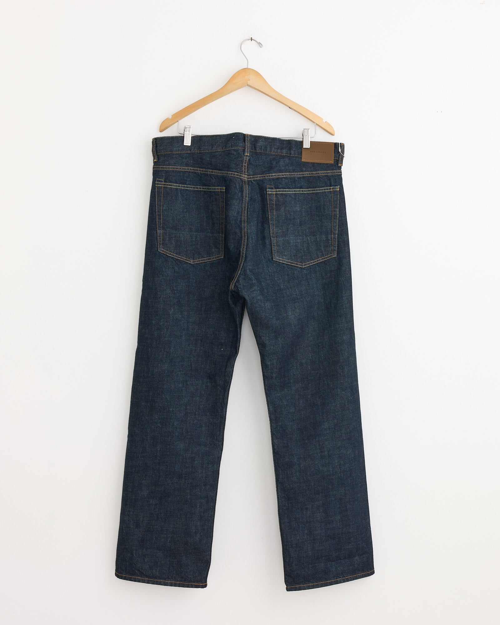 A pair of Our Legacy Third Cut Jeans in Blue Selvedge hangs from a wooden hanger against a white wall, shown from the back with visible pockets and stitching in premium Italian chain twill.