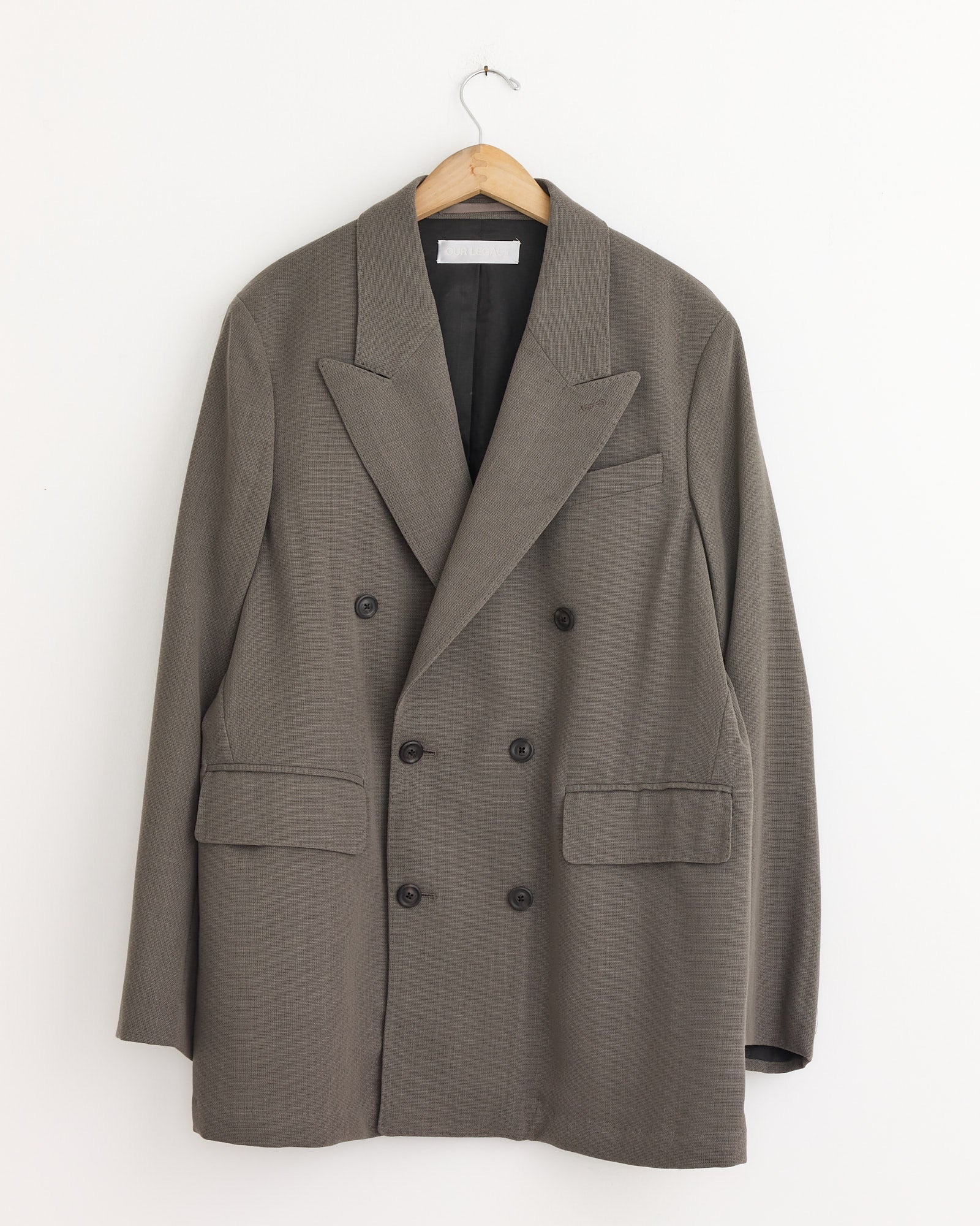 The Sharp DB Blazer in Double Grey by Our Legacy, a men’s double-breasted blazer with wide lapels and flap pockets, hangs on a wooden hanger against a white wall.
