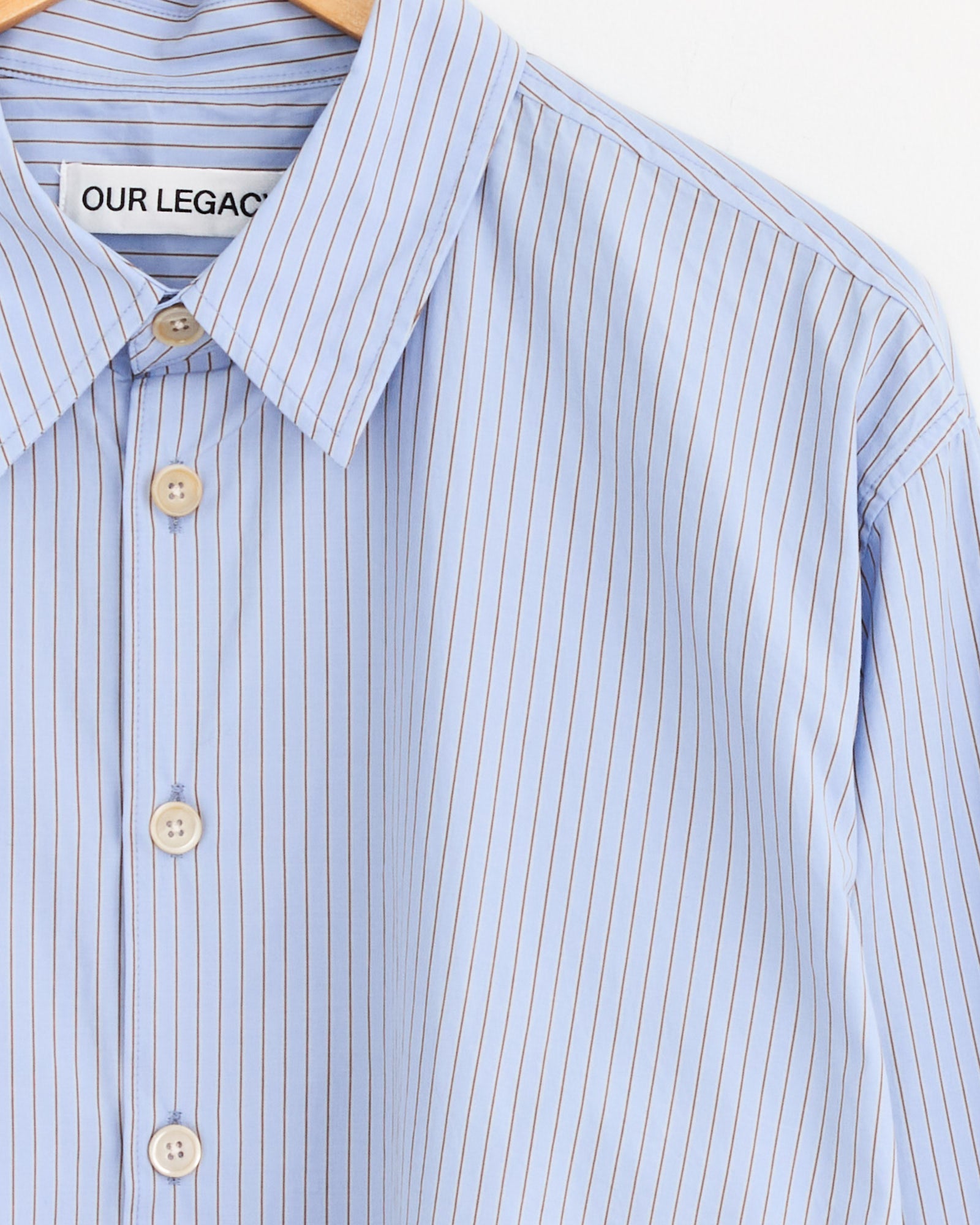 The Beyond Shirt in Typewriter Stripe by Our Legacy is a light blue Japanese cotton button-up with thin white and brown stripes, white buttons, and a collar label. It hangs on a wooden hanger against a white background.