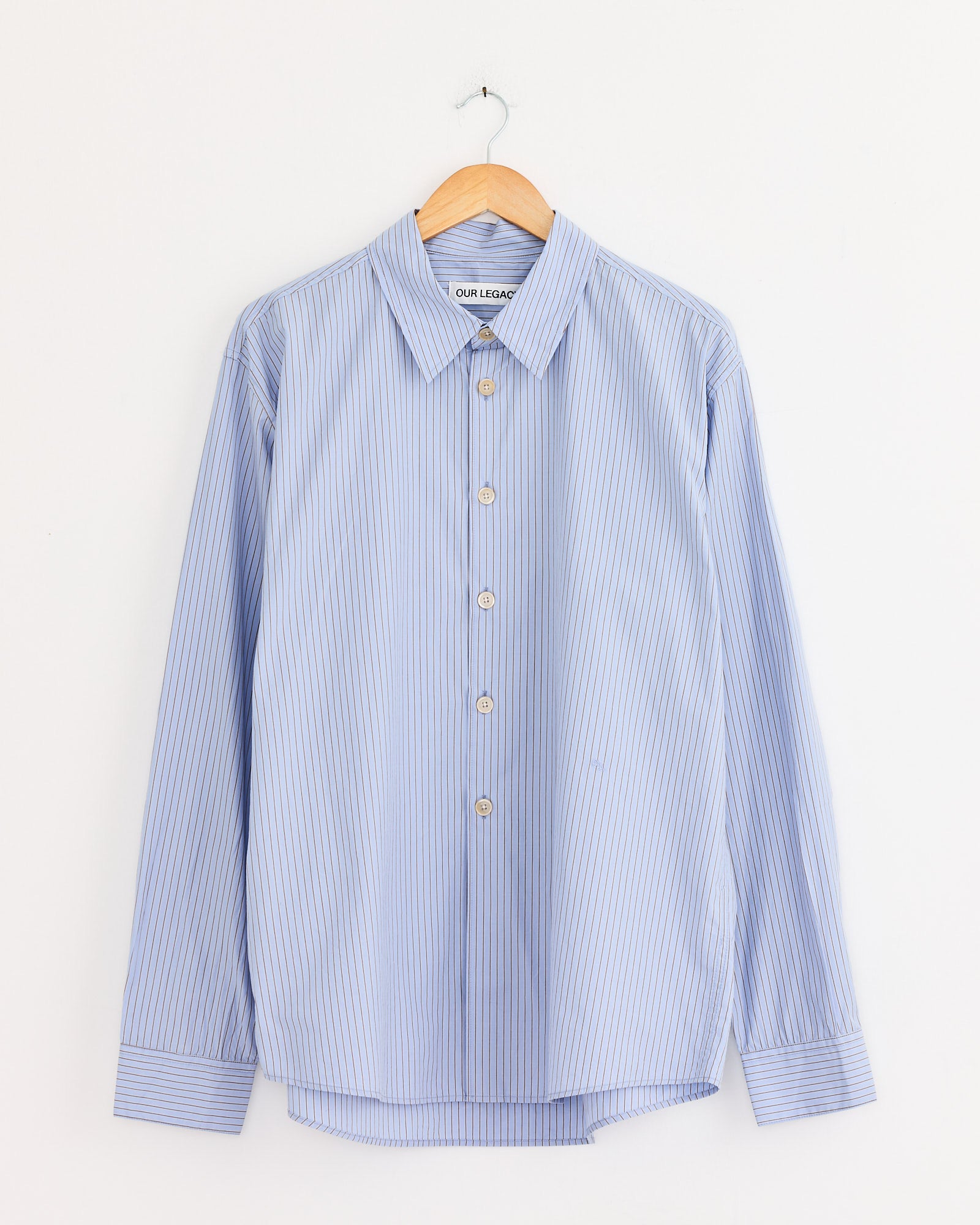 The Our Legacy Beyond Shirt in Typewriter Stripe, a light blue Japanese cotton long-sleeve button-up with thin white stripes, hangs on a wooden hanger against a plain white wall.
