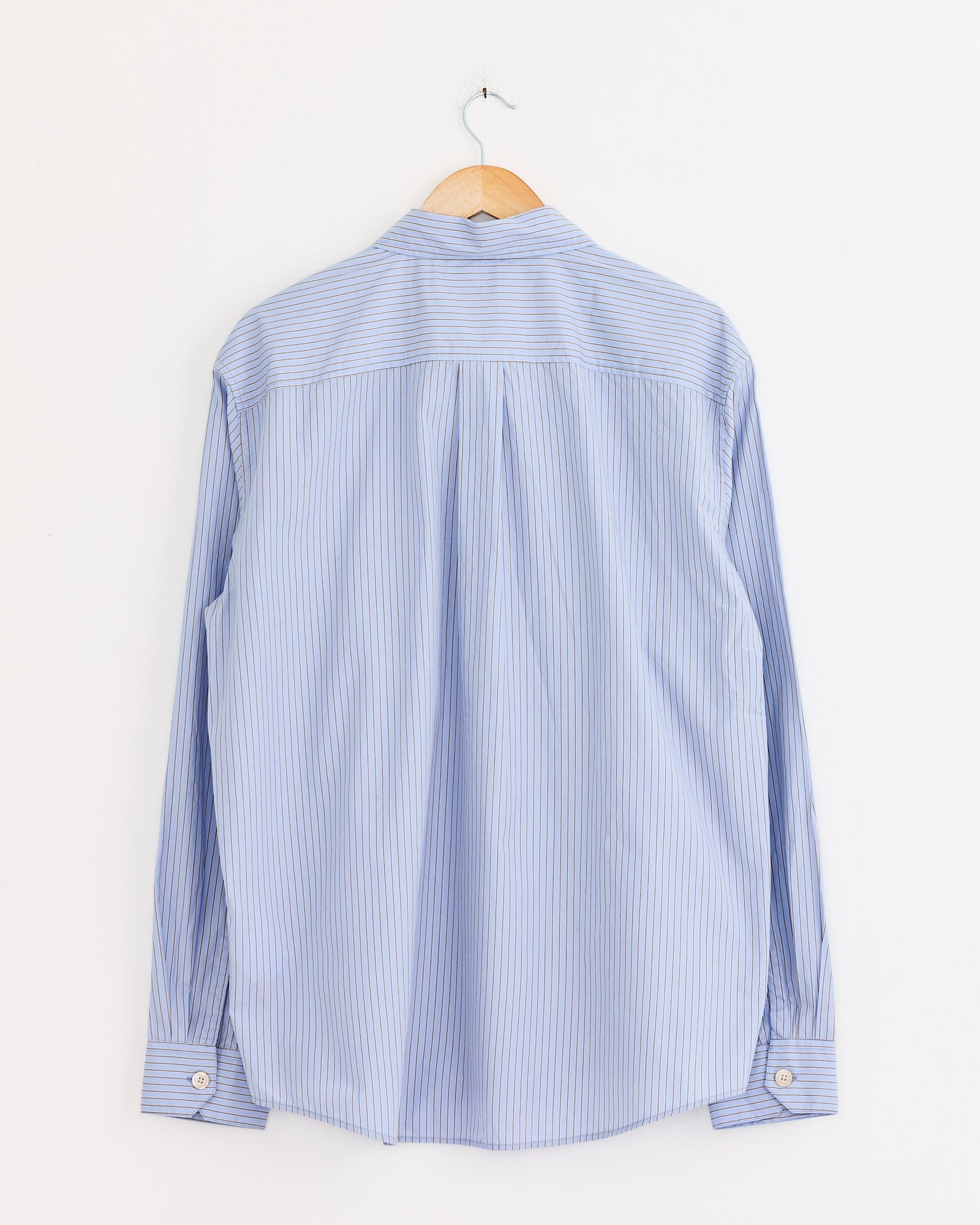 The Our Legacy Beyond Shirt in Typewriter Stripe, a light blue long-sleeve shirt with thin white stripes, made from Japanese cotton poplin, hangs on a wooden hanger against a plain white wall, shown from the back.