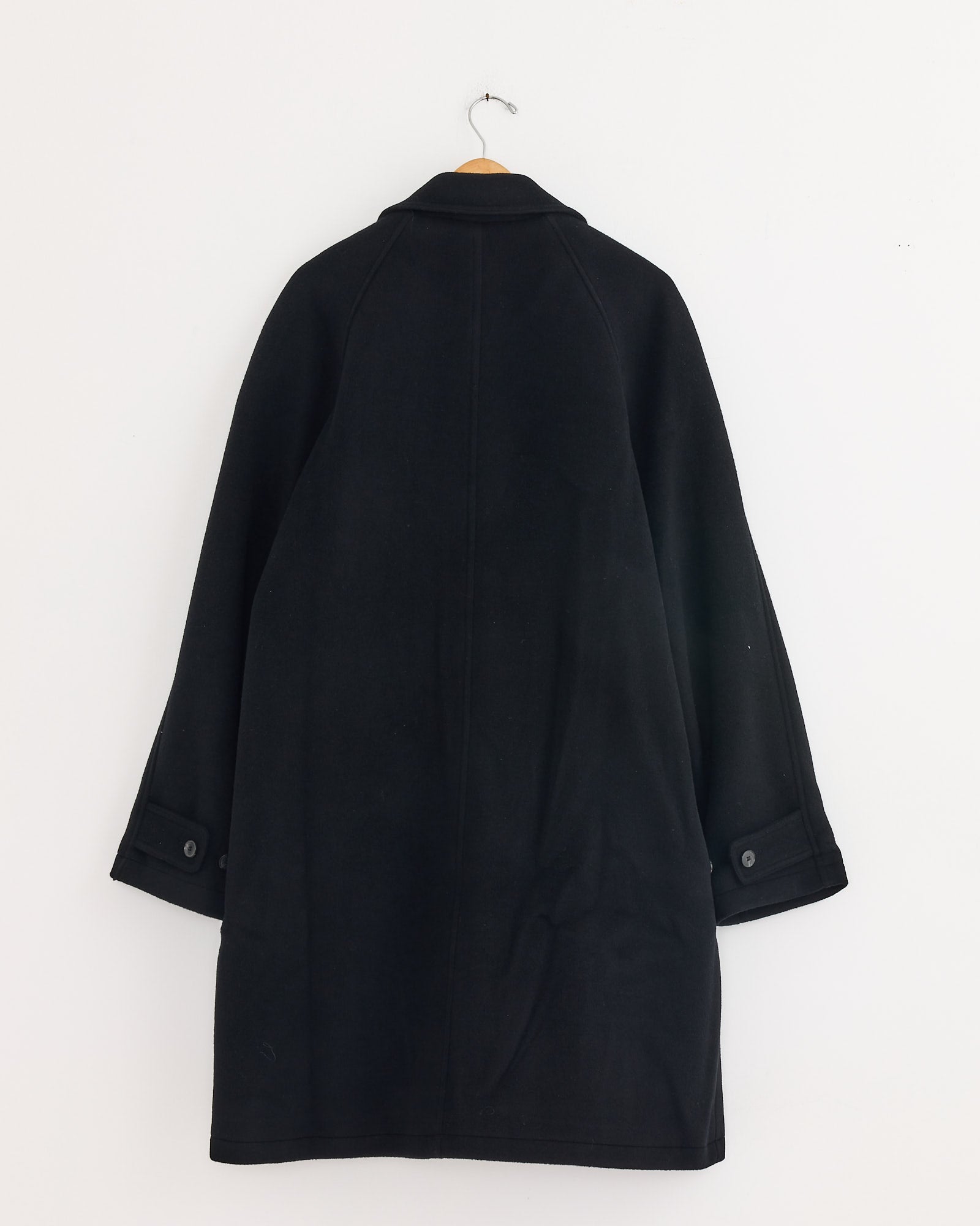 The mfpen Accession Coat in Black, featuring long sleeves and buttoned cuffs, is displayed on a wooden hanger against a plain white wall with its back facing forward.