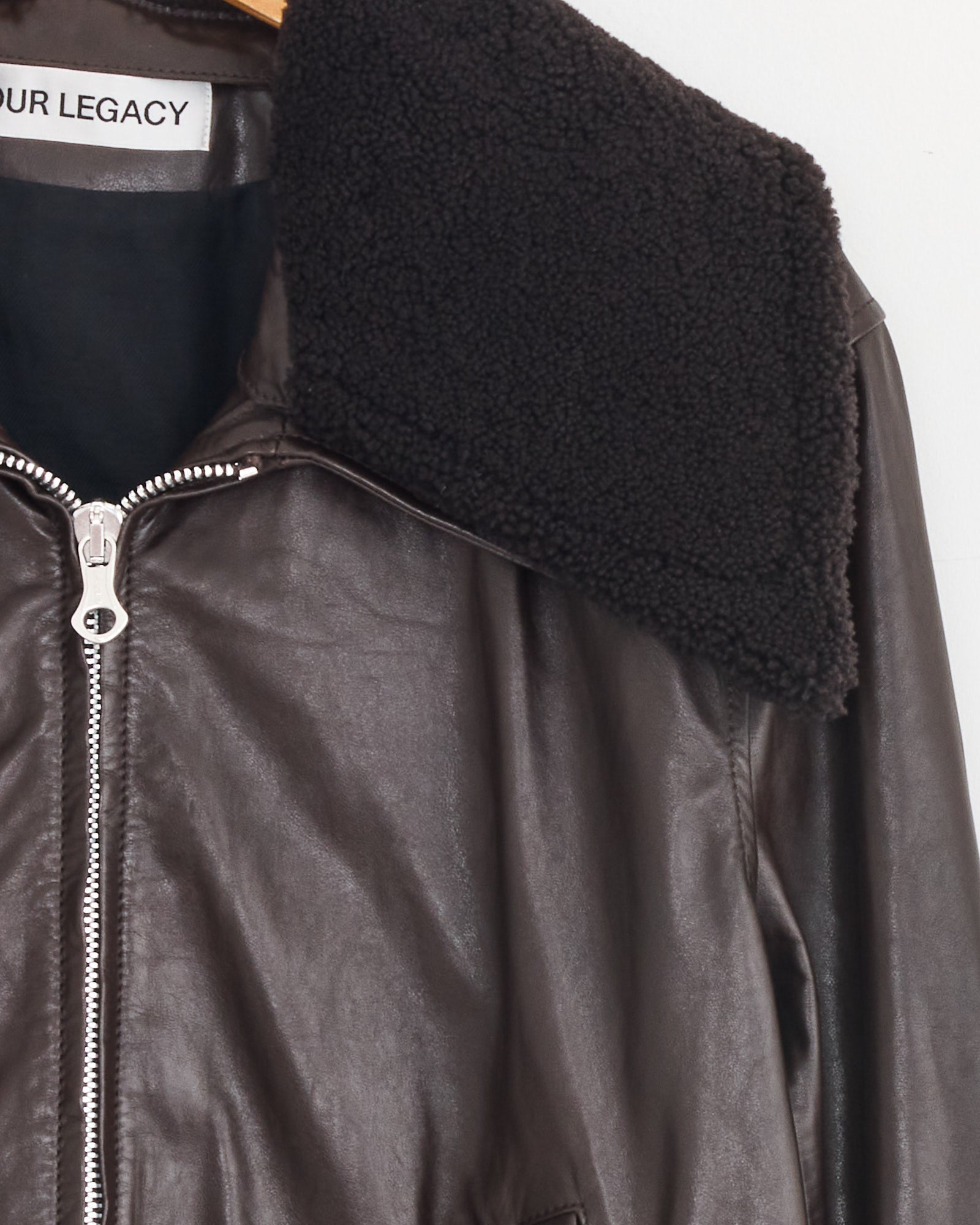 Close-up of the Our Legacy Pharos Bomber Jacket in Umber Brown, crafted from calfskin leather with a textured shearling collar and silver zipper. The jacket hangs on a wooden hanger, label "OUR LEGACY" inside the collar.