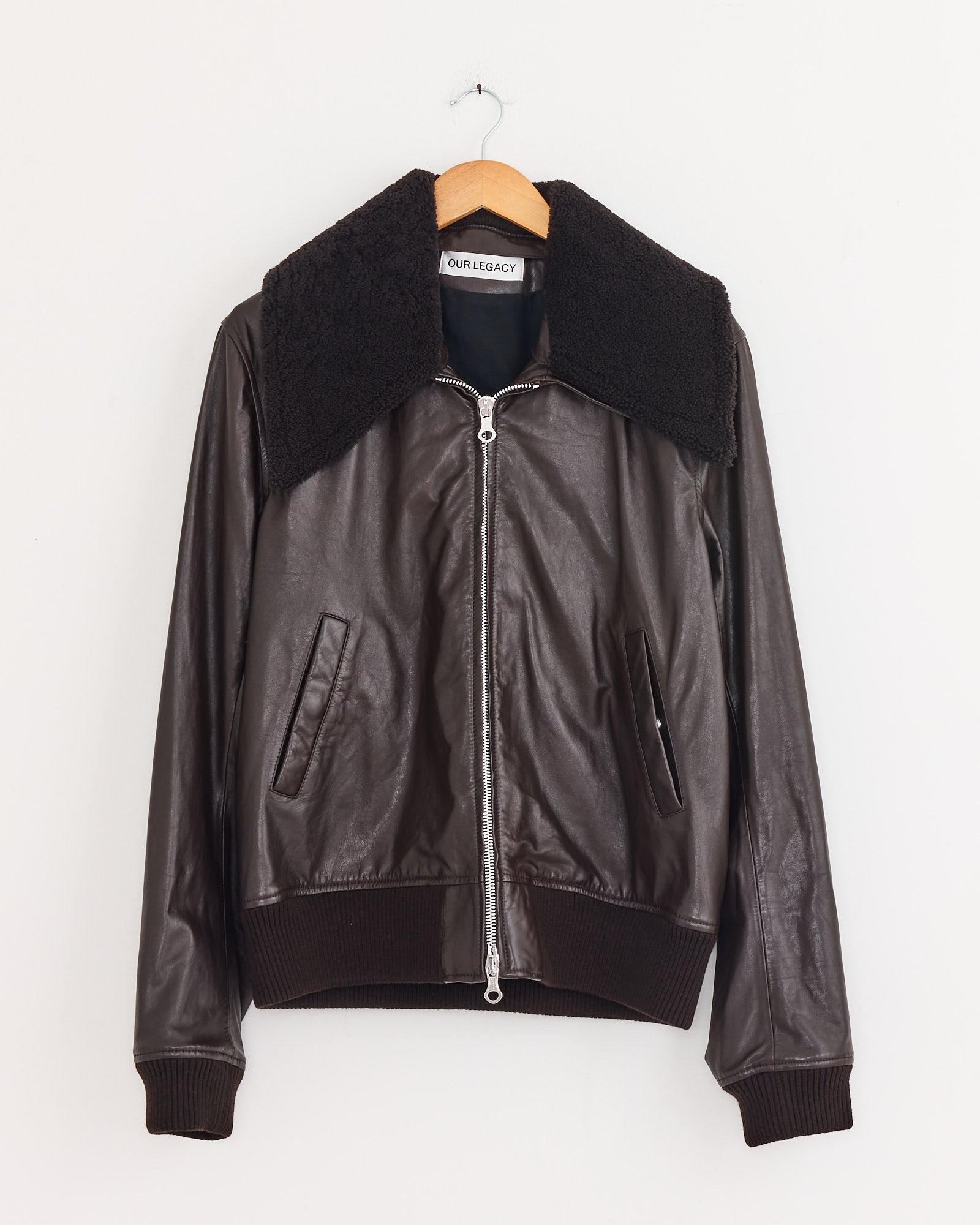 The Our Legacy Pharos Bomber Jacket in Umber Brown, featuring calfskin leather, a black shearling collar, front zipper, ribbed cuffs and hem, and two slanted side pockets, hangs on a wooden hanger against a plain white background.