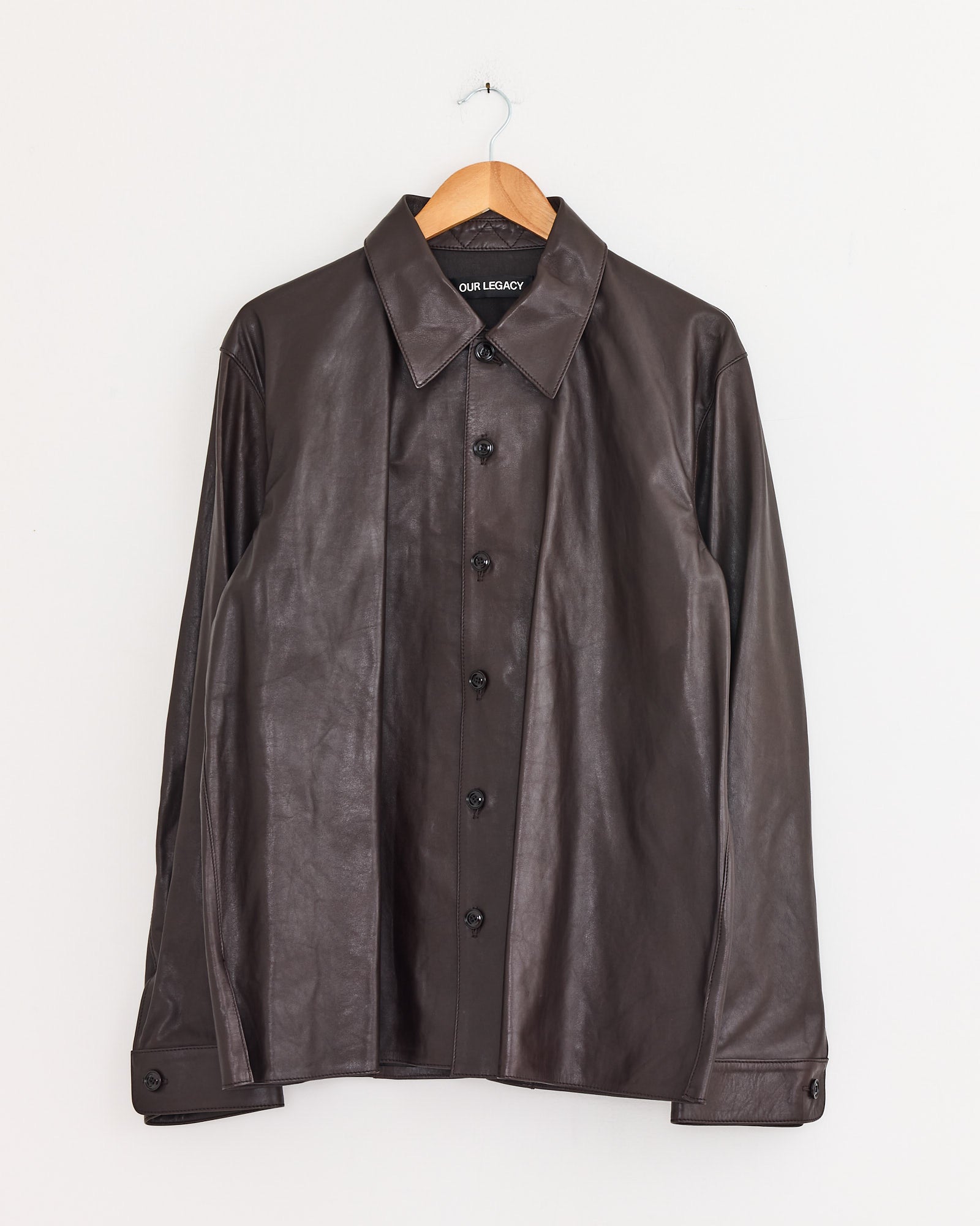 The Our Legacy Welding Shirt in Umber Brown calfskin leather, featuring a collar and button-up design, hangs on a wooden hanger against a plain white wall with the brand label visible inside the collar.