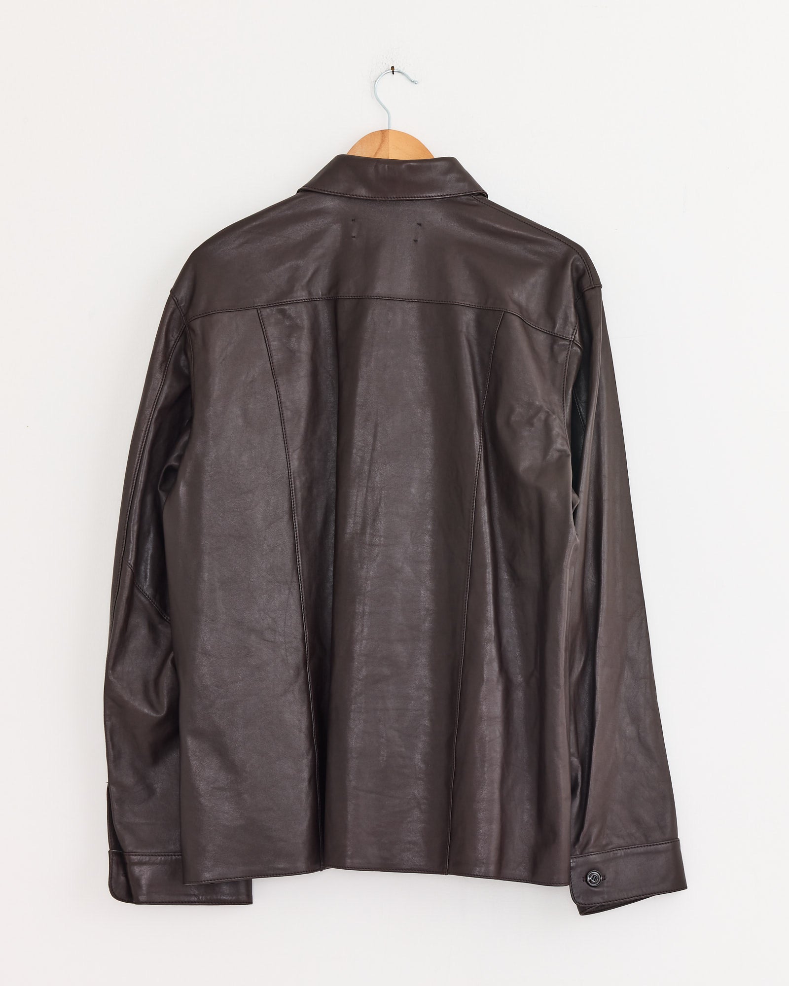 The Our Legacy Welding Shirt in Umber Brown calfskin hangs from a wooden hanger against a white wall, shown from the back with long sleeves and a visible collar, echoing the brand’s classic style.