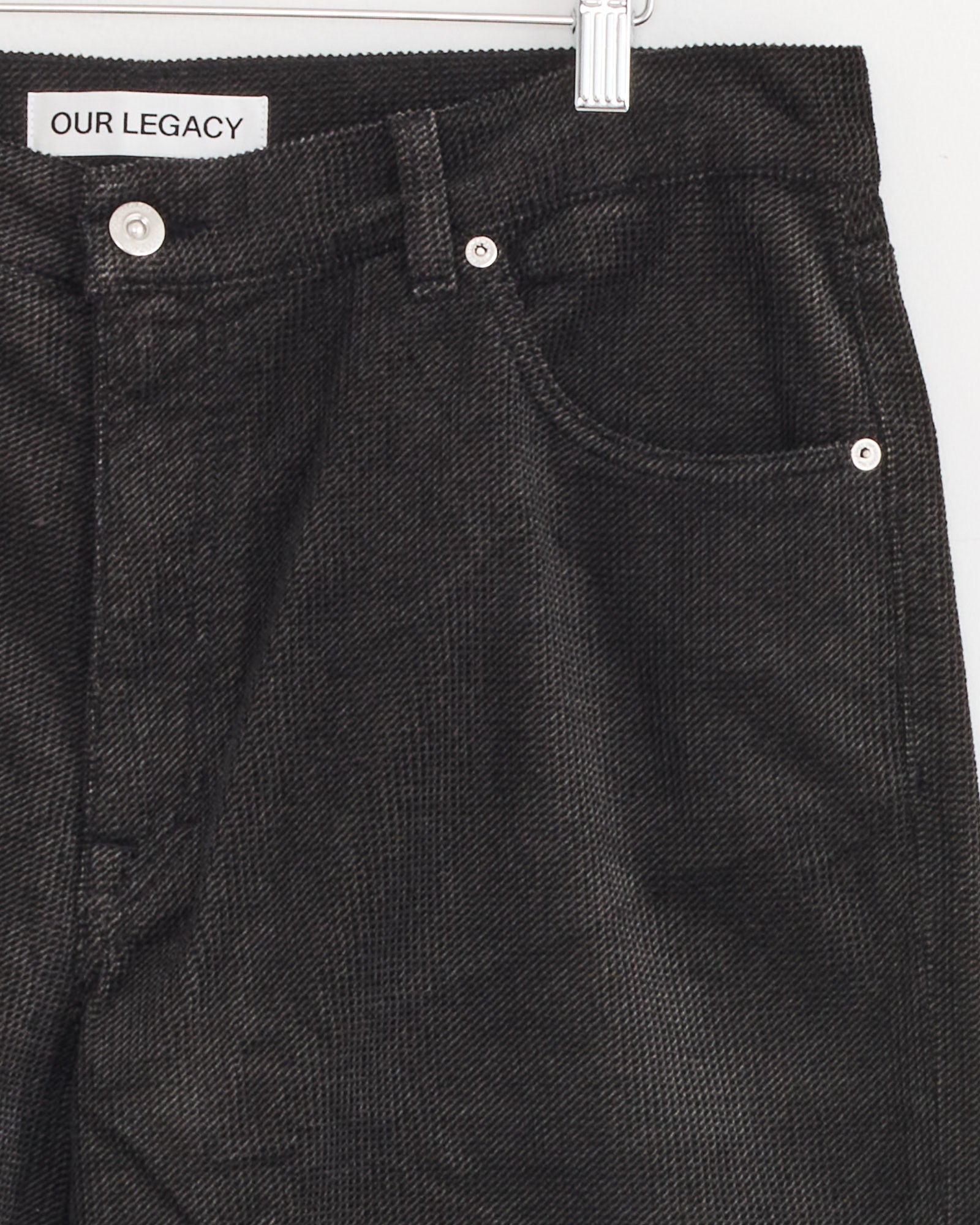 Close-up view of the front pocket and waistband of Our Legacy Vast Cut Corduroy Pants in dark brown, featuring a visible inner label. The textured corduroy fabric adds depth, and the pants are displayed on a metal hanger.