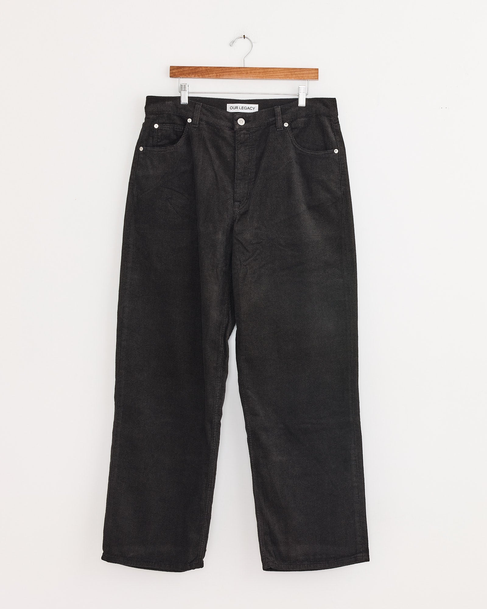 A pair of Our Legacy Vast Cut Corduroy Pants in dark brown hangs on a wooden hanger against a plain white wall.