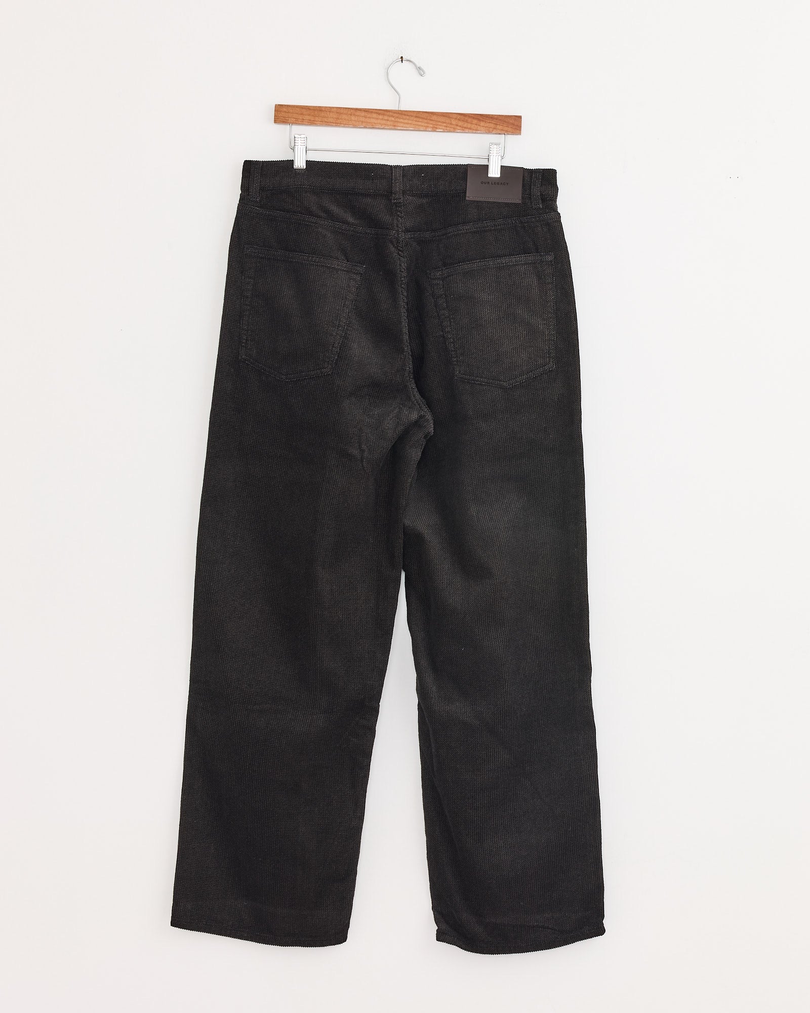 A pair of Our Legacy Vast Cut Corduroy Pants in dark brown with a wide loose fit, hanging on a wooden hanger against a plain white wall, shown from the back with visible pockets and straight legs.
