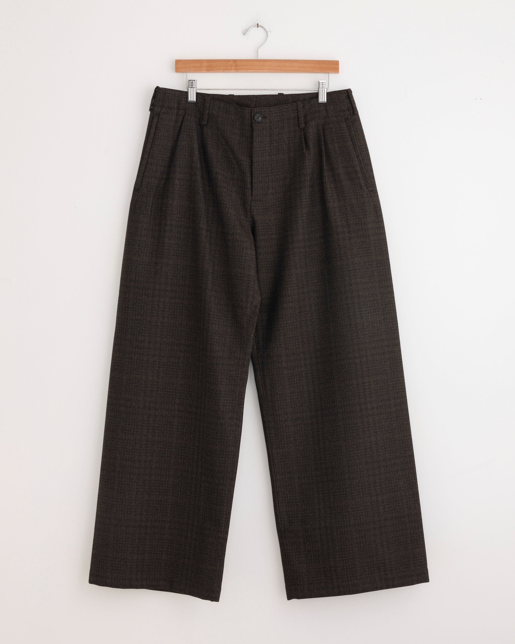 The Bridge Chino in Brown by Our Legacy, featuring a wide-leg plaid design in Italian virgin wool, hangs on a wooden hanger against a plain white wall.