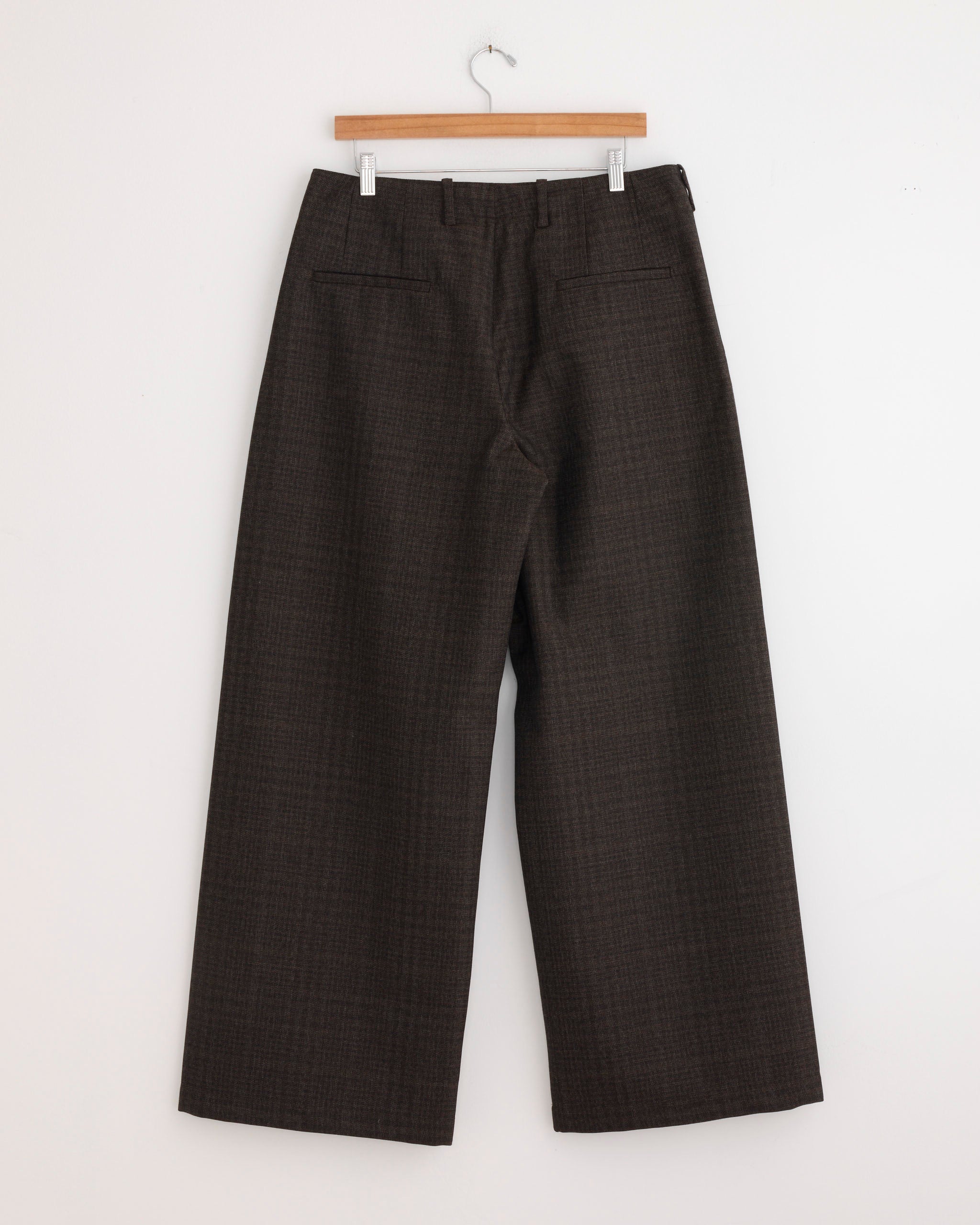 The Bridge Chino in Brown by Our Legacy, crafted from Italian virgin wool with a subtle check pattern, features a wide-leg silhouette, high waist, and back welt pockets. Shown hanging on a wooden hanger against a white wall.