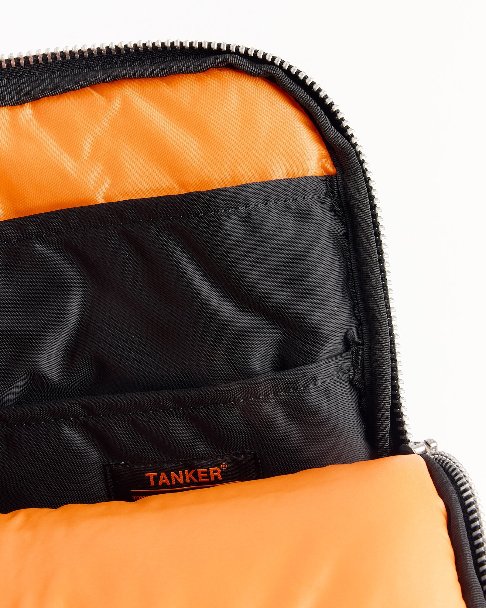 Close-up of the Porter Tanker Document Case in Black: open bag with silver zipper, bright orange lining, black inner pockets, and a black label with orange "TANKER" text visible inside.