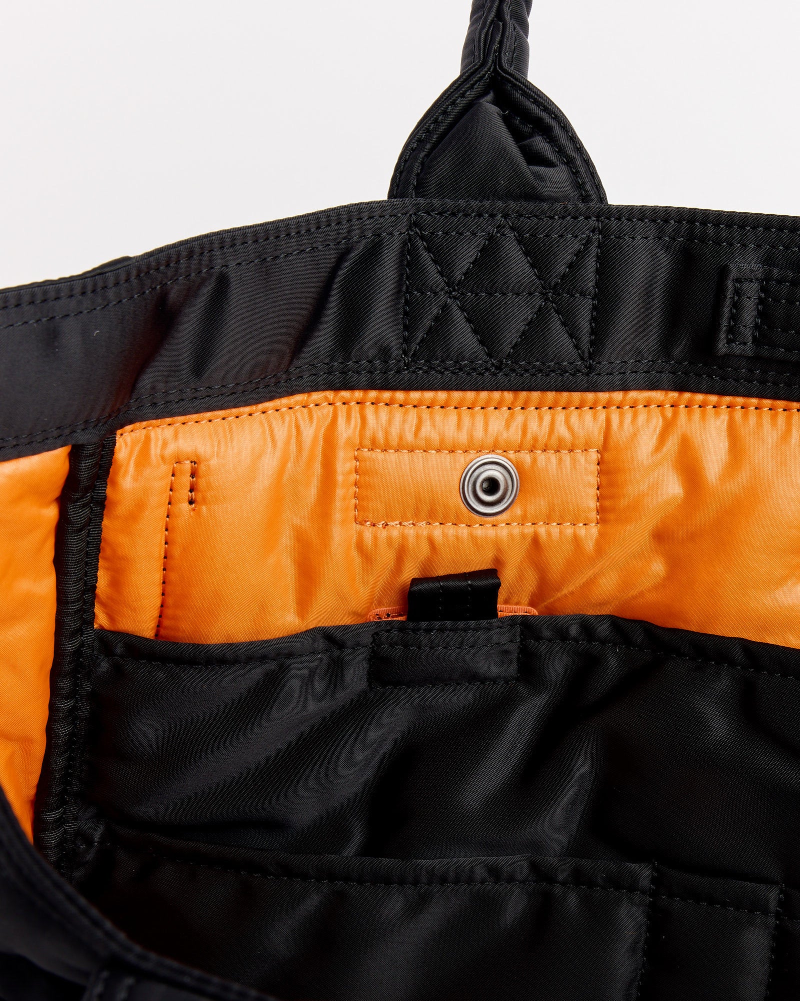 Close-up of the Porter Tanker Tote Bag in Black, featuring an orange lining, multiple pockets, a snap button closure, visible stitching, and a fabric loop.