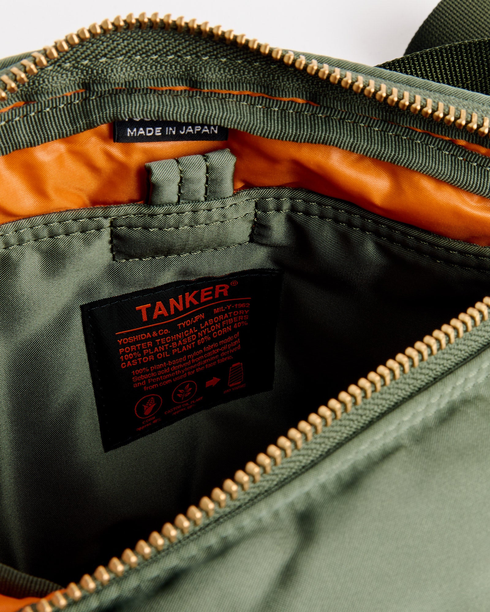 Close-up of an open Porter Tanker Fanny Pack in Sage Green with a gold zipper, orange lining, black TANKER® label with product info, and a small MADE IN JAPAN tag near the zipper.