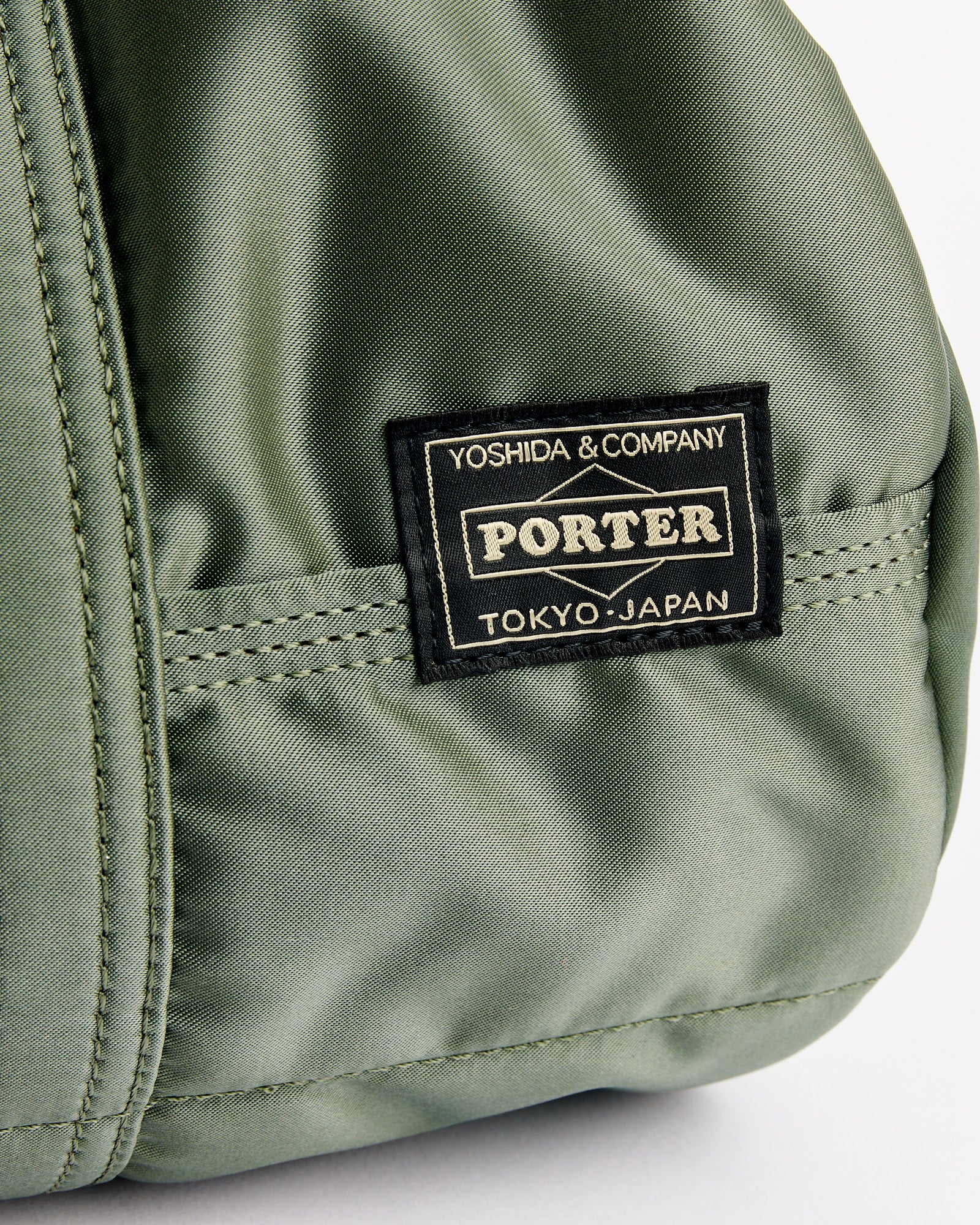 Close-up of the Tanker Roll Bag in Sage Green by Porter, featuring a stitched black label reading "Yoshida & Company Porter Tokyo, Japan.