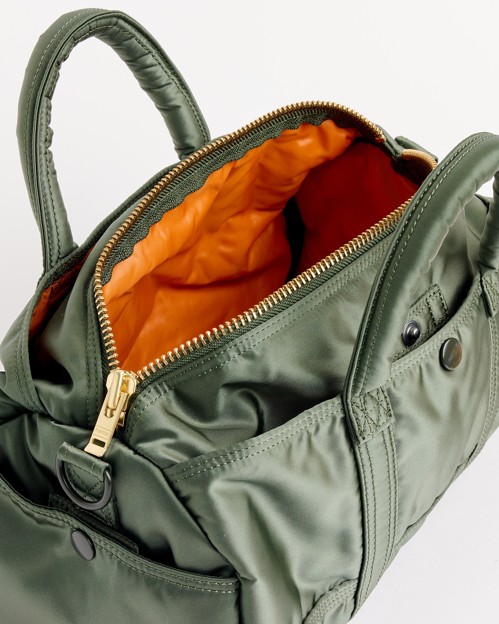 The Porter Tanker Roll Bag in Sage Green features sturdy handles, exterior snap button pockets, a gold zipper, and opens to reveal a vibrant orange lining.