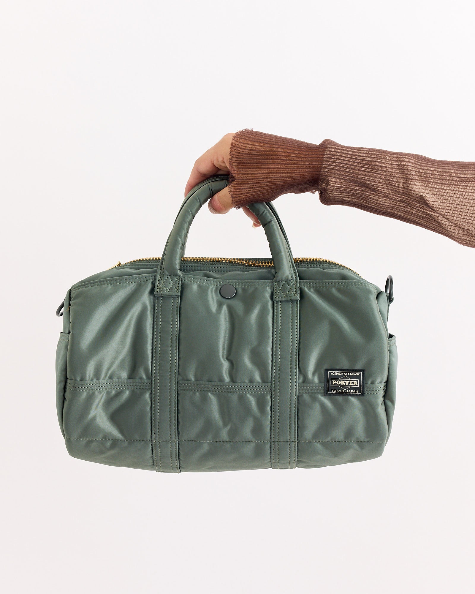 A hand in a brown ribbed sleeve holds the Porter Tanker Roll Bag in sage green, featuring nylon material, two handles, front pockets, and a black rectangular label, set against a plain white background.