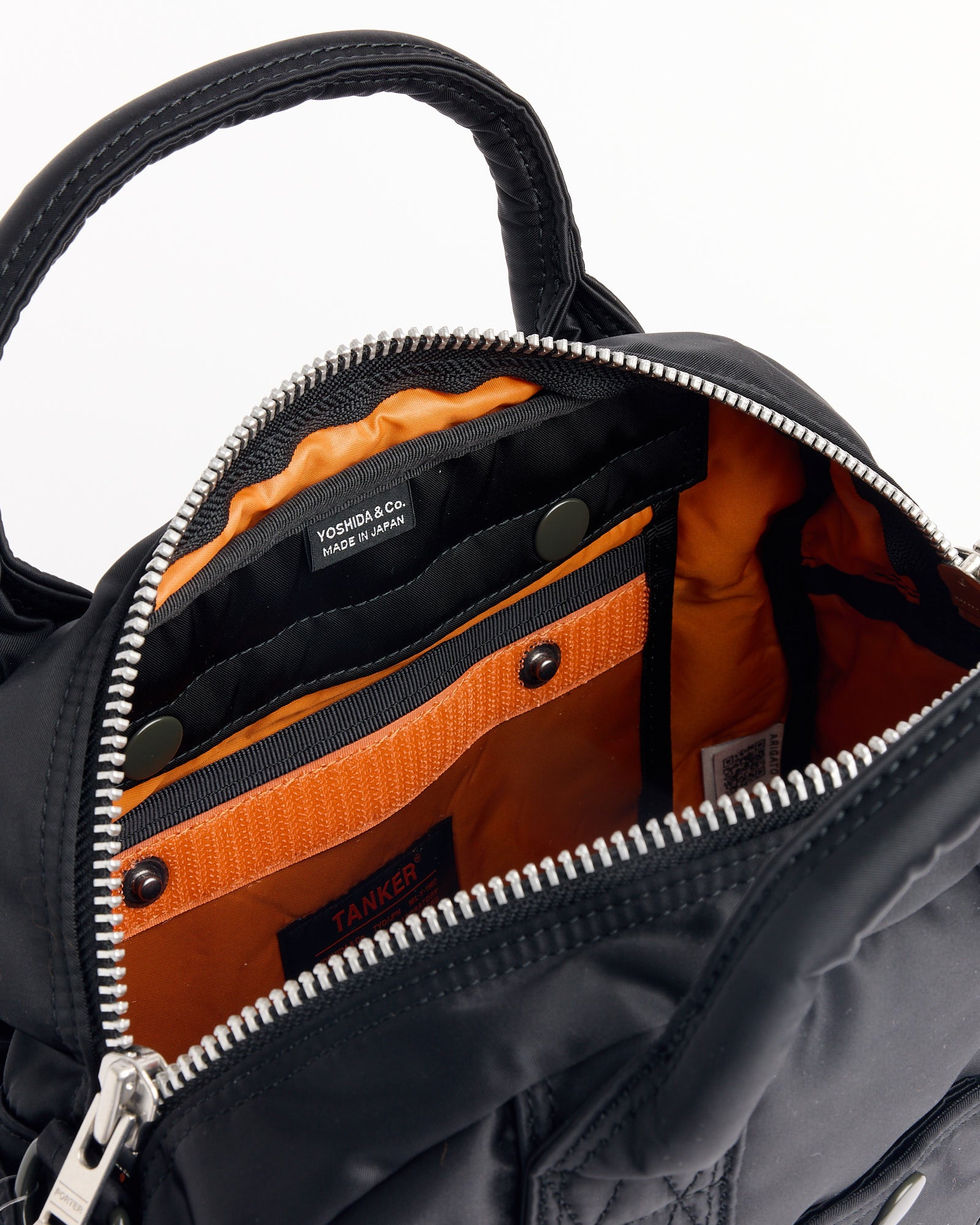A close-up of the Porter Tanker Boston Bag in Black displays its silver zipper, bright orange interior, multiple pockets, and a Yoshida & Co. Made in Japan label inside.