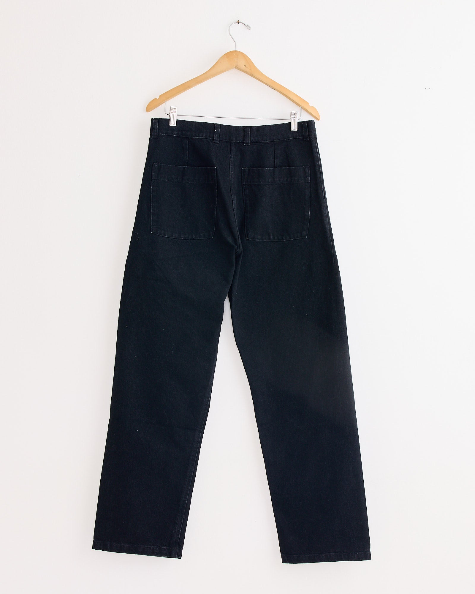 The Arpenteur 5 Pocket Trouser in Black features straight legs and two large back pockets, shown on a wooden hanger against a white wall.
