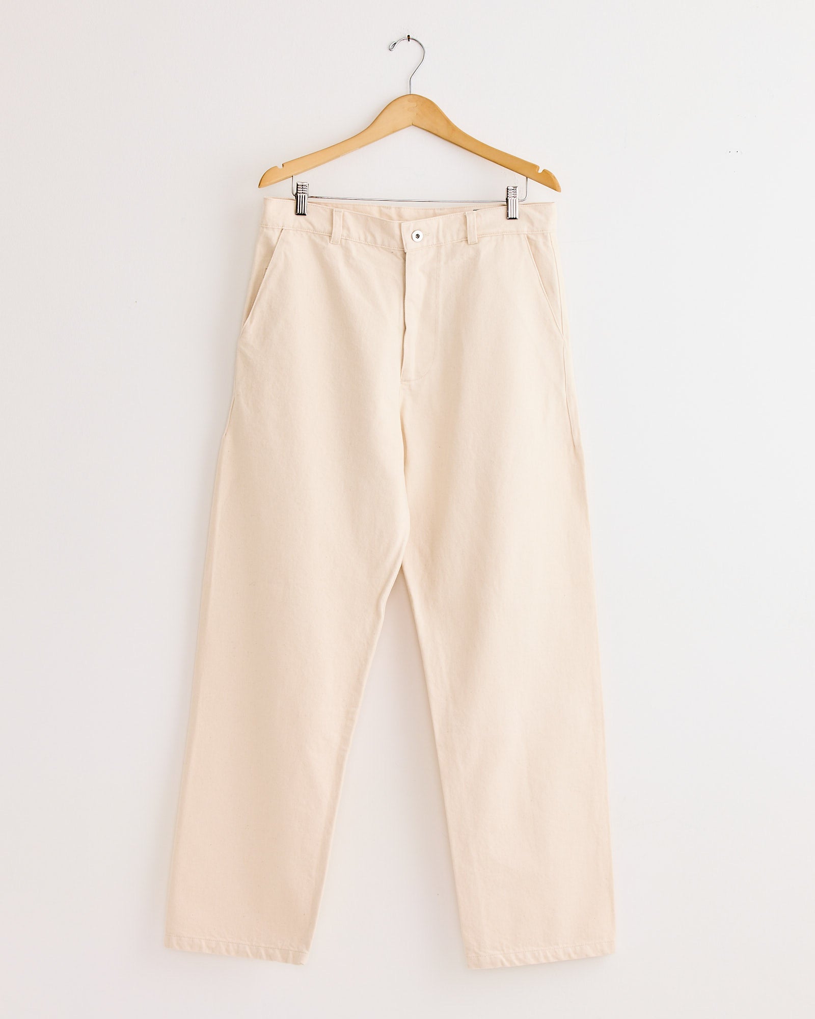 The Arpenteur 5 Pocket Trouser in Ecru, a high-waisted straight-leg style, hangs on a wooden hanger against a plain white wall.