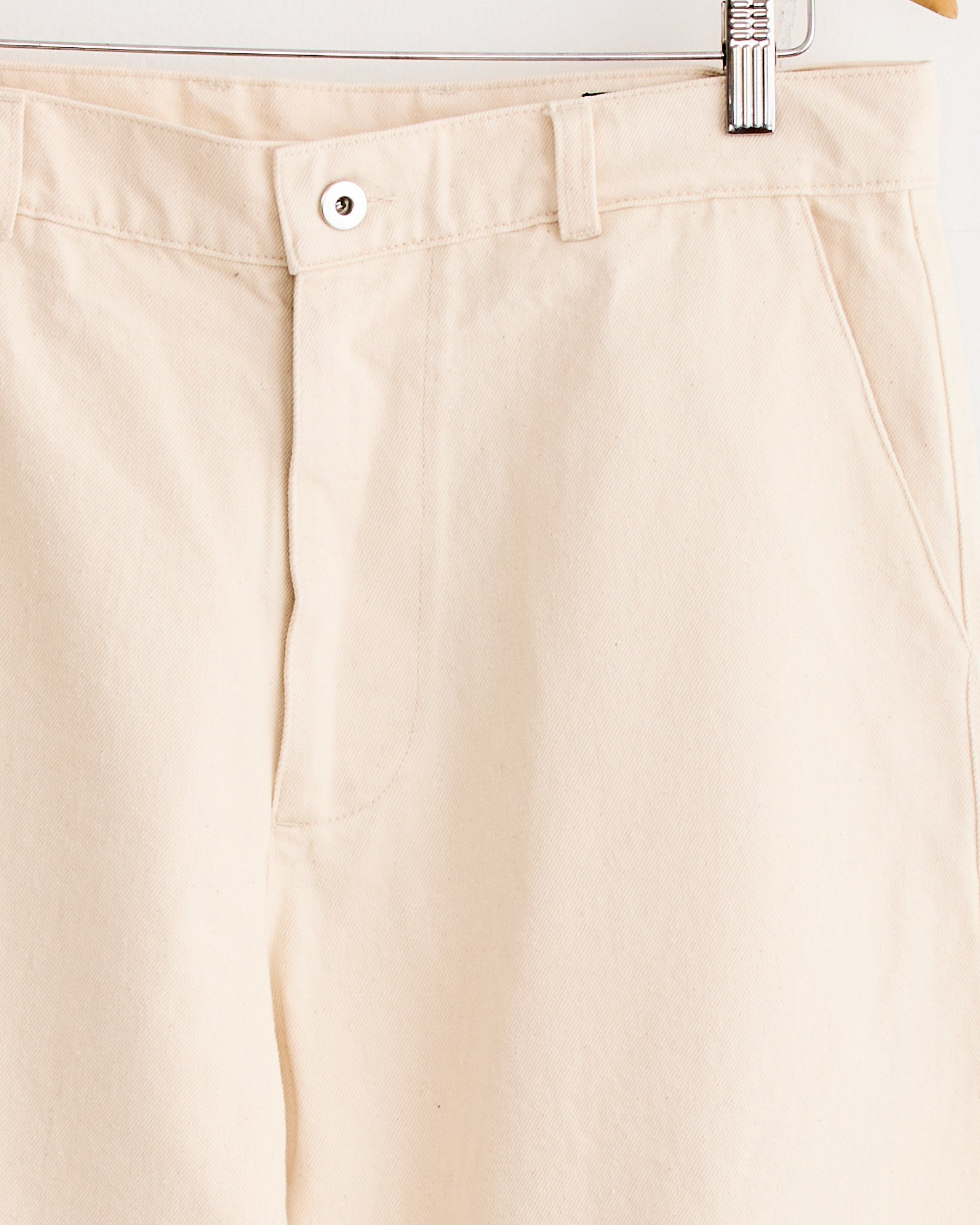 Close-up of Arpenteur's 5 Pocket Trouser in Ecru, crafted from indigo cotton twill in France, shown on a hanger with visible waistband, button, belt loops, pockets, and zipper top against a light background.