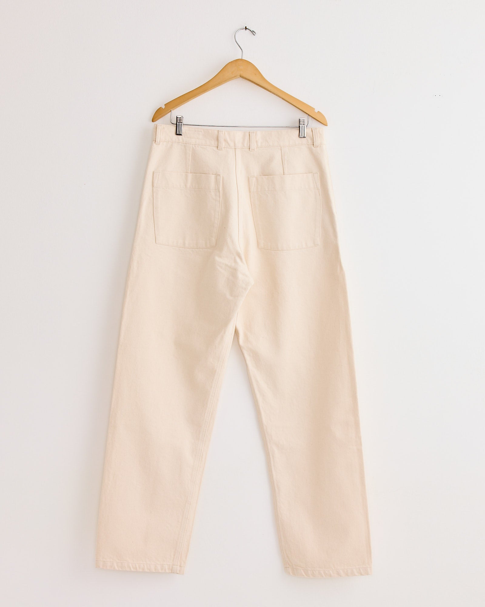 The Arpenteur 5 Pocket Trouser in Ecru hangs on a wooden hanger against a white wall, displaying the back with two large square pockets and straight legs.