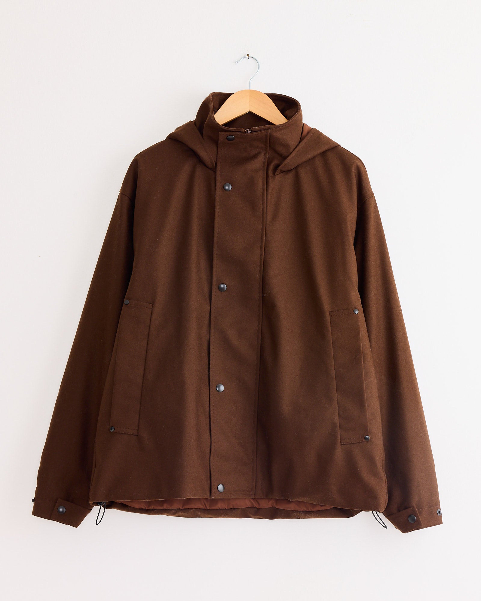 The Arpenteur Air Primaloft Filled Parka Coat in Tobacco, featuring snap buttons and two front pockets, hangs on a wooden hanger against a plain white wall.