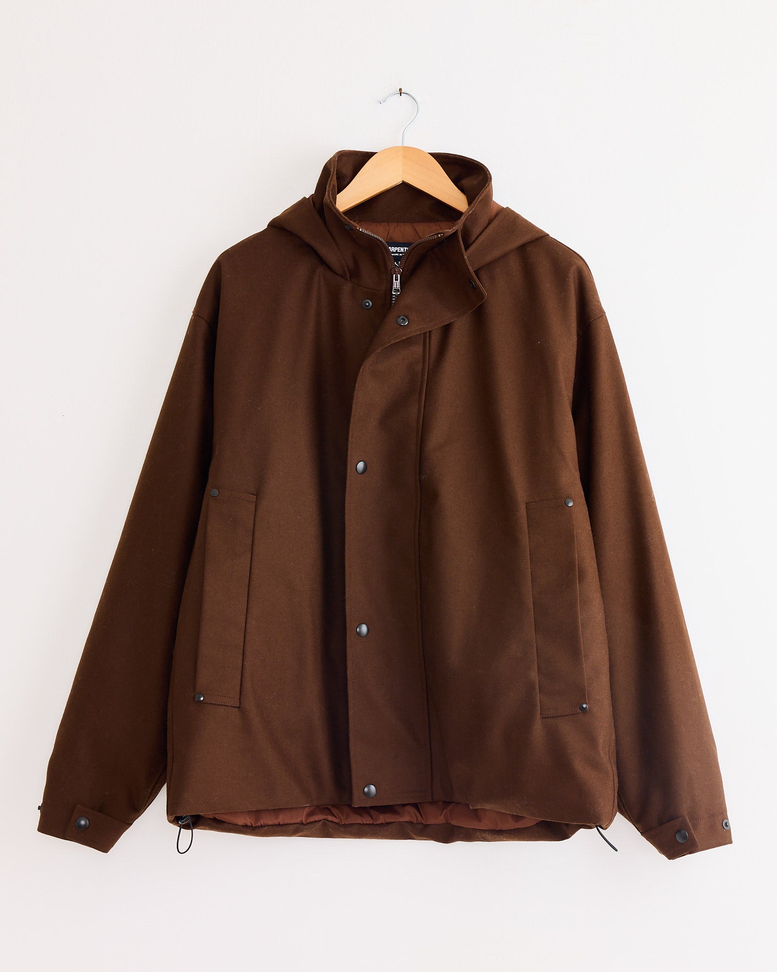 The Arpenteur Air Primaloft Filled Parka Coat in Tobacco, featuring snap buttons and two front pockets, hangs on a wooden hanger against a white wall.