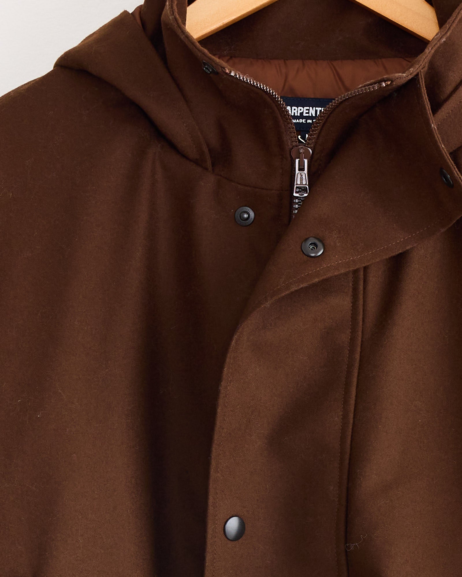A close-up of the Arpenteur Air Primaloft Filled Parka Coat in Tobacco on a wooden hanger, showing black buttons and a partially visible zipper beneath the collar.