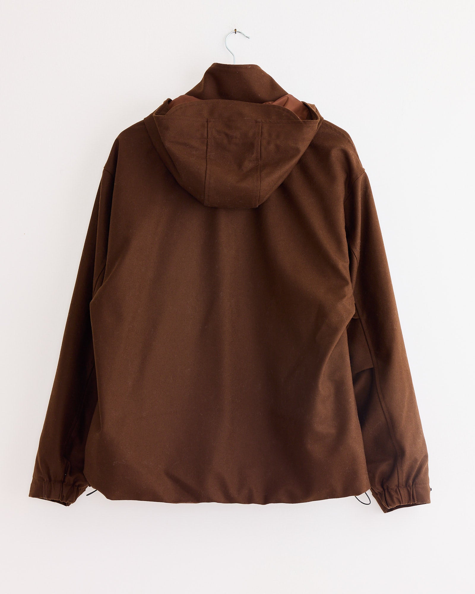 An Arpenteur Air Primaloft Filled Parka Coat in Tobacco hangs from a simple metal hook against a white wall, displayed from the back.