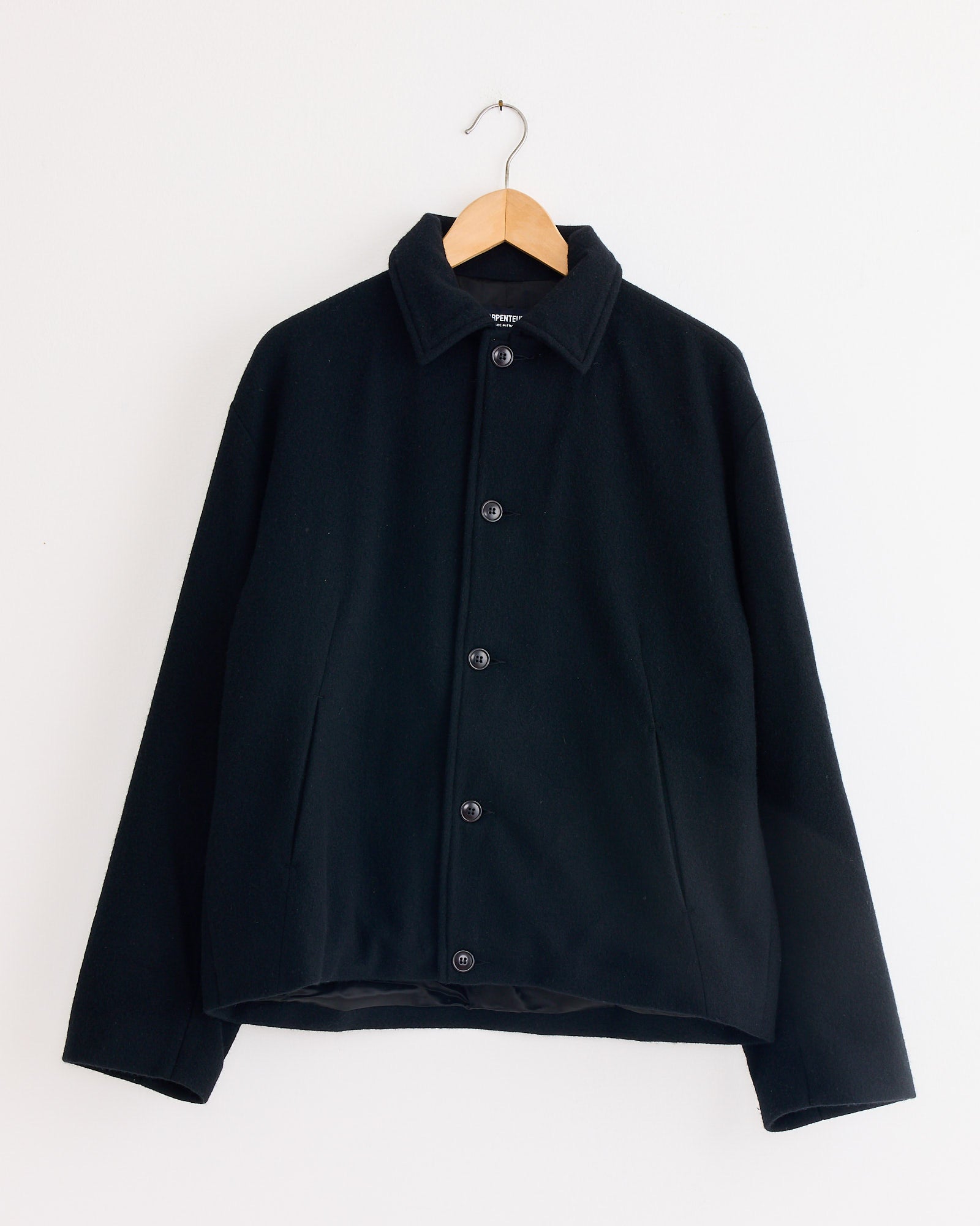 The Arpenteur Lux J Lined Jacket in Black, a collared button-up workwear piece, hangs on a wooden hanger against a plain white wall.