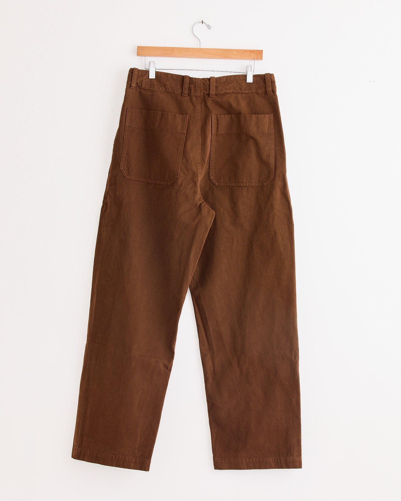 The Arpenteur Lux P Trouser in Tobacco, crafted in France, hangs on a wooden hanger against a white wall, displayed from the back with visible belt loops and two large back pockets.