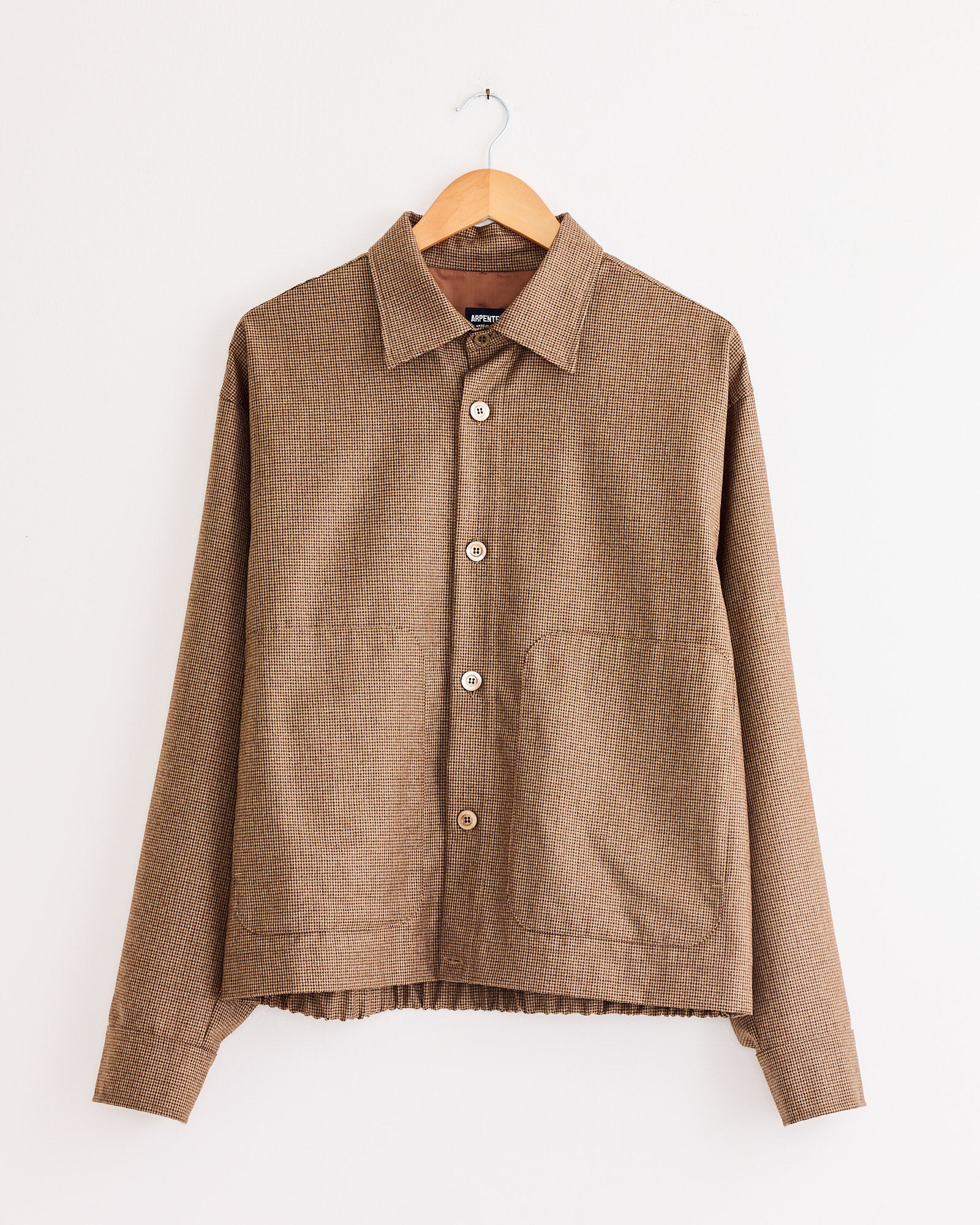 The Arpenteur Sigma Overshirt Jacket in Houndstooth is a brown, long-sleeve, 100% wool jacket with a button-up front, collar, and large front pockets, shown on a wooden hanger against a plain white wall.