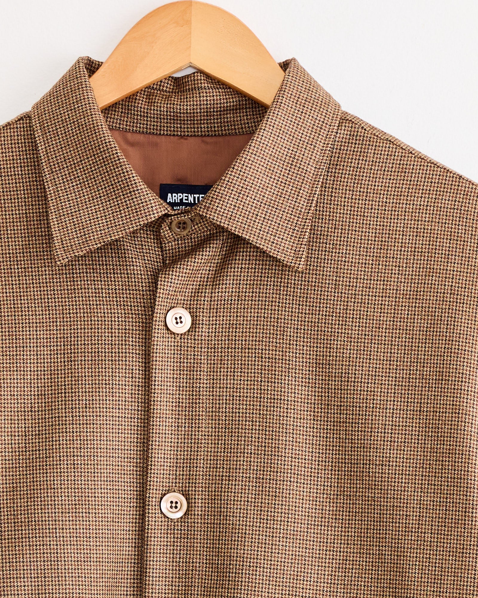 The Arpenteur Sigma Overshirt Jacket in brown and beige houndstooth features a pointed collar, white buttons, and a visible brand label. Crafted from 100% wool, it combines comfort and style.