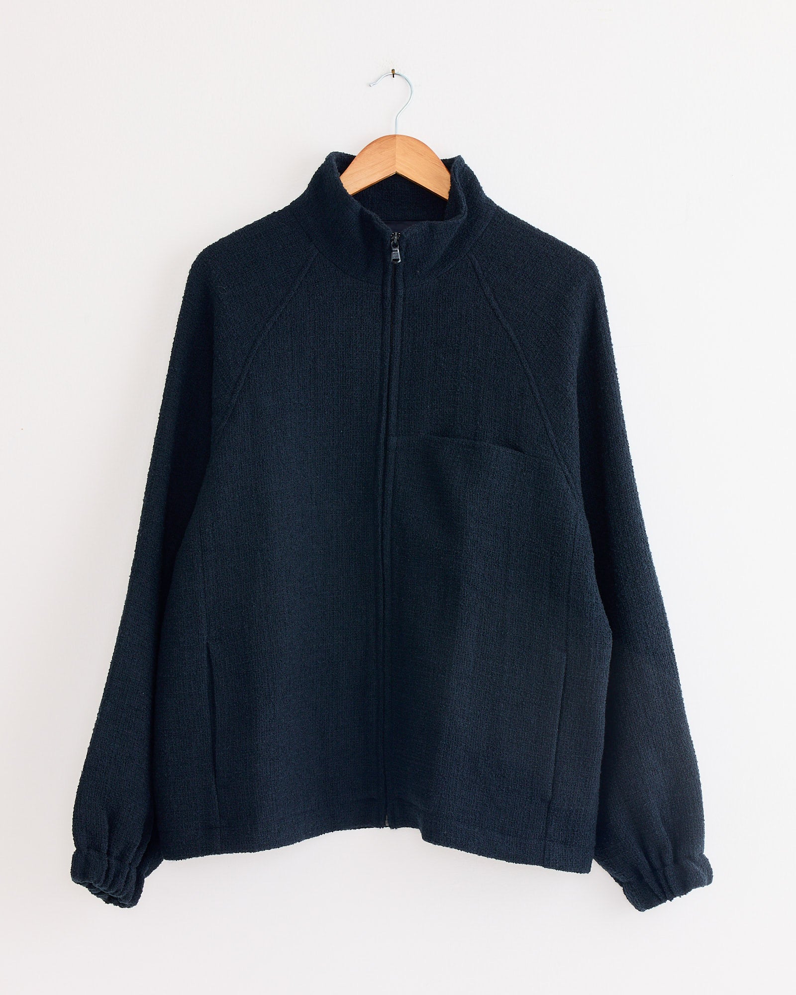 The Arpenteur Track J Zippered Jacket in Midnight, featuring a high collar, long sleeves, and two front pockets, hangs on a wooden hanger against a plain white wall.