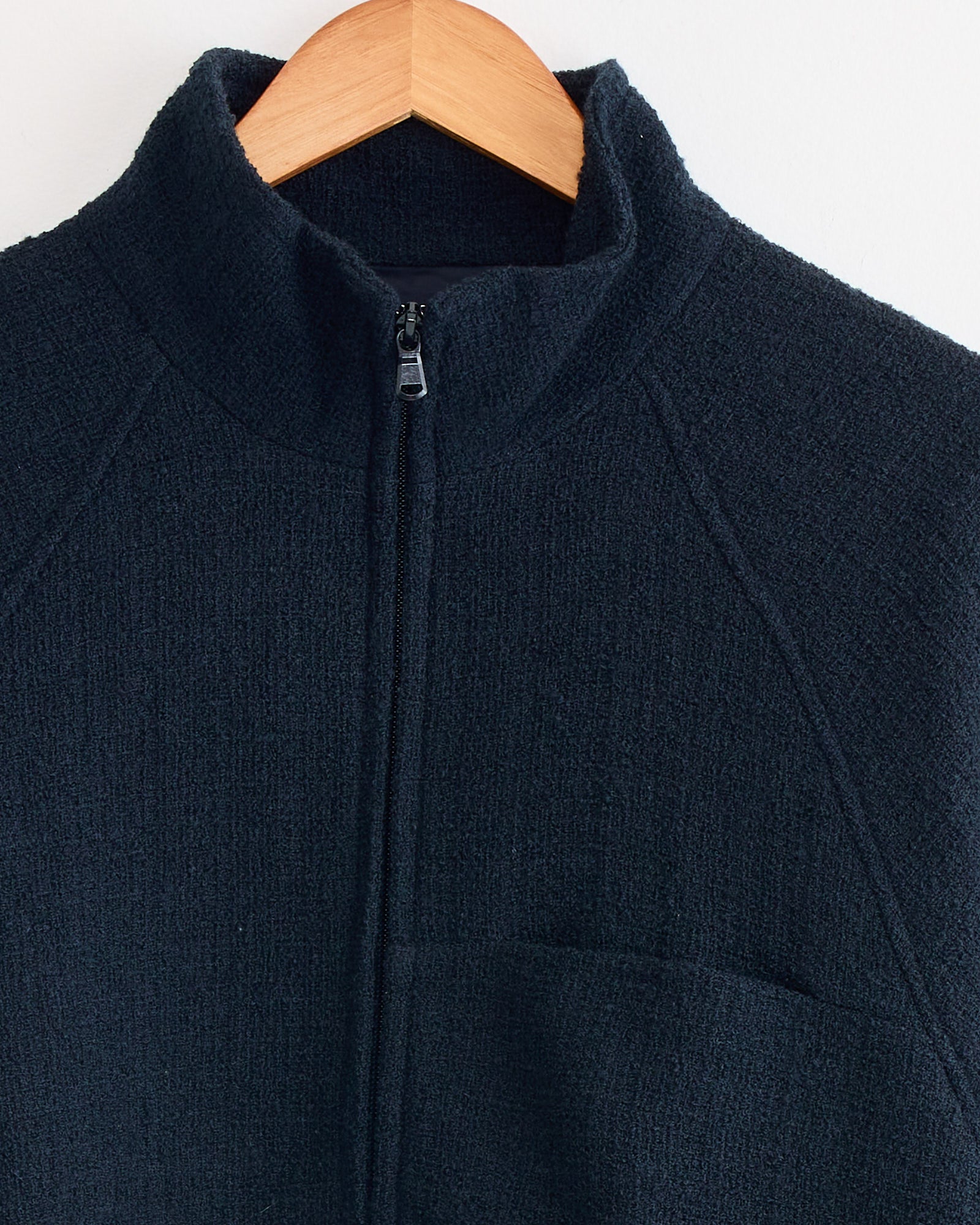 A close-up of the Arpenteur Track J Zippered Jacket in Midnight shows the collar, partial zipper, and upper chest pocket as it hangs on a wooden hanger against a white background.