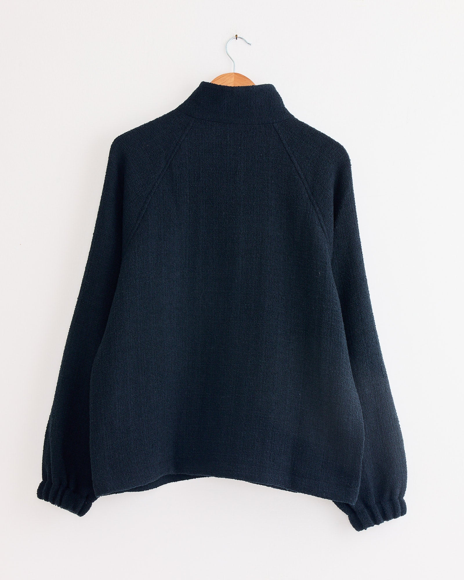 The Arpenteur Track J Zippered Jacket in Midnight, a loose-fit, long-sleeved woolen tweed jacket with elastic cuffs and high collar, hangs on a wooden hanger against a plain white wall. Made in France.