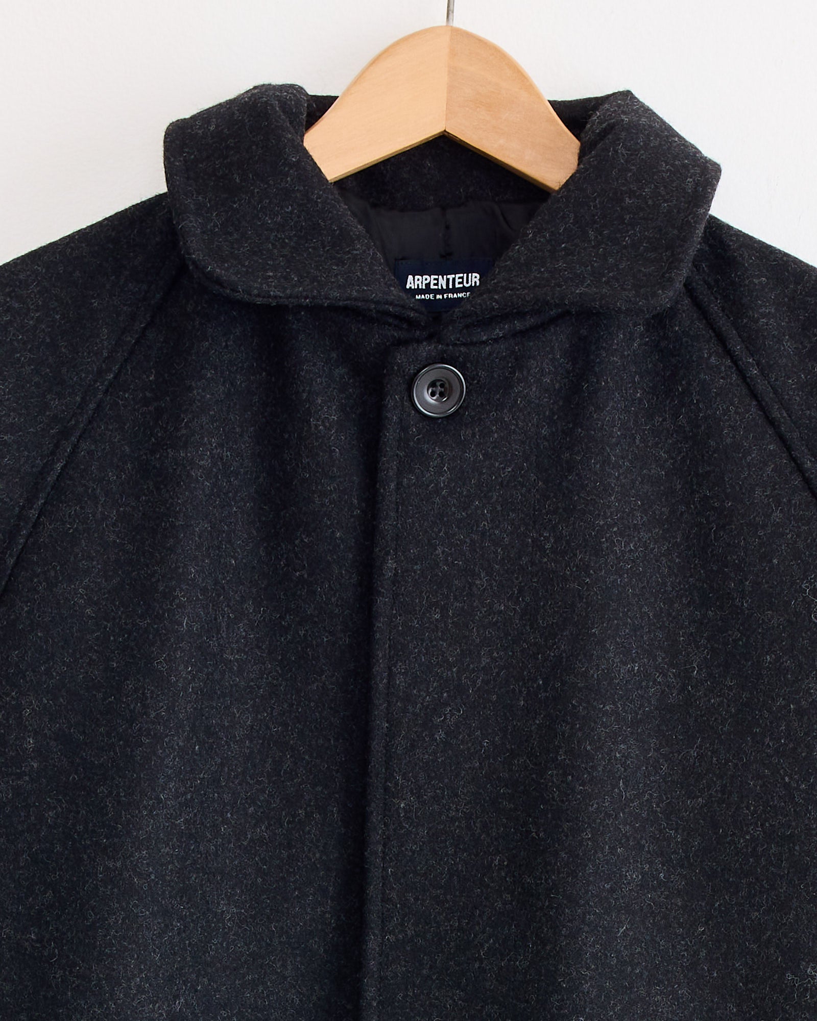 The Arpenteur Utile Lined Wool Coat in Charcoal hangs on a wooden hanger against a light background, featuring a single visible button at the collar. Made in France, as indicated on the inner label.