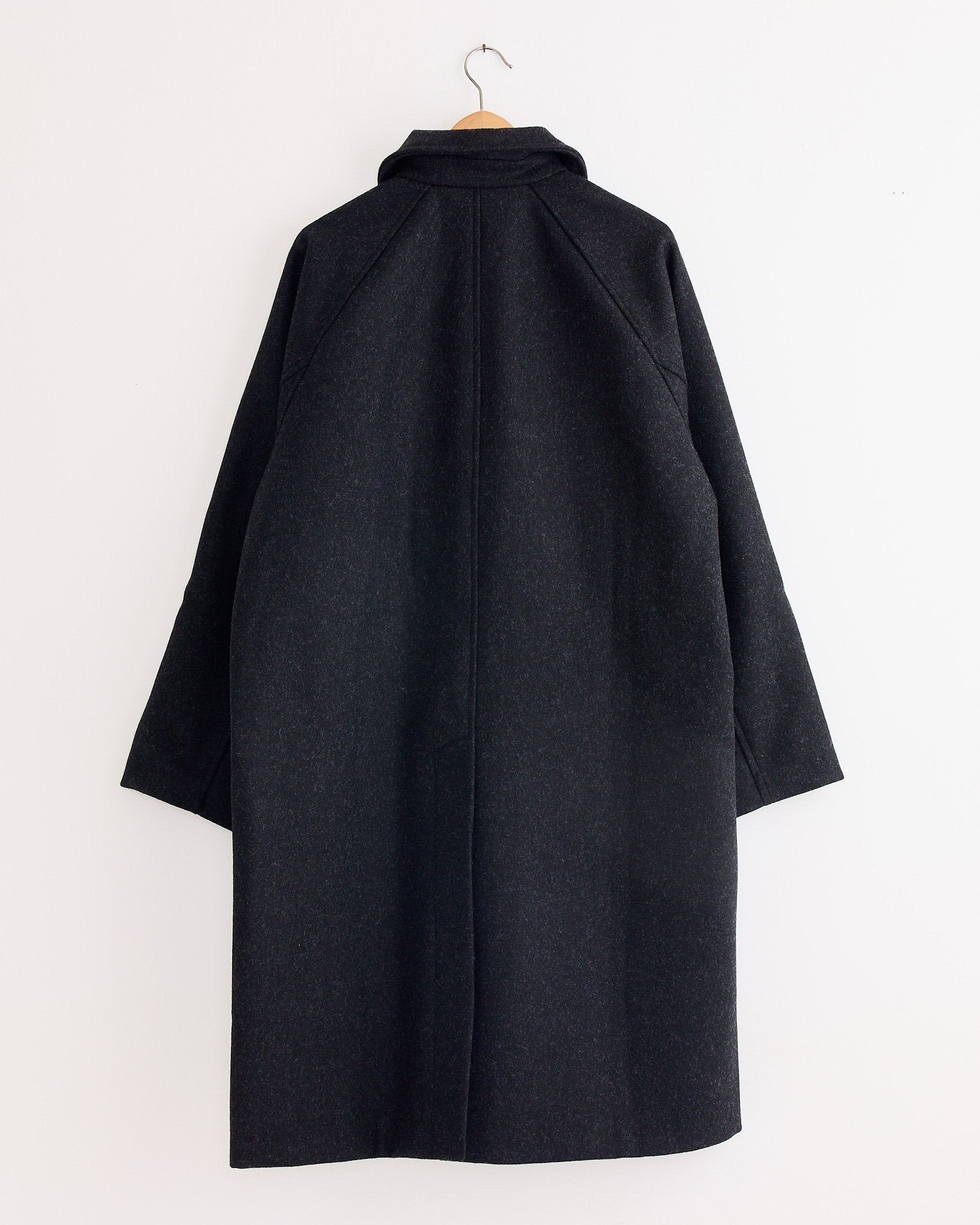 The Arpenteur Utile Lined Wool Coat in Charcoal, made in France, hangs on a wooden hanger against a white wall, shown from the back with wide sleeves and a high collar.
